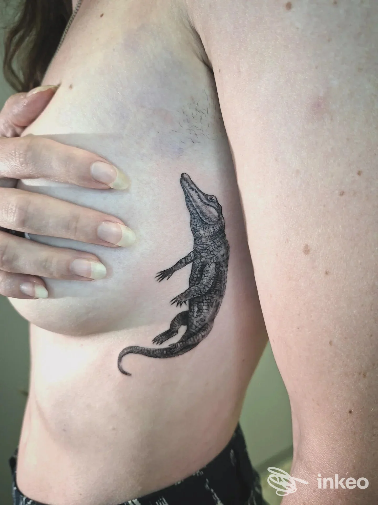 Tattoo realistico di un lucertola situato sul fianco sinistro di una persona, visibile la pelle e una mano che copre parzialmente la zona.