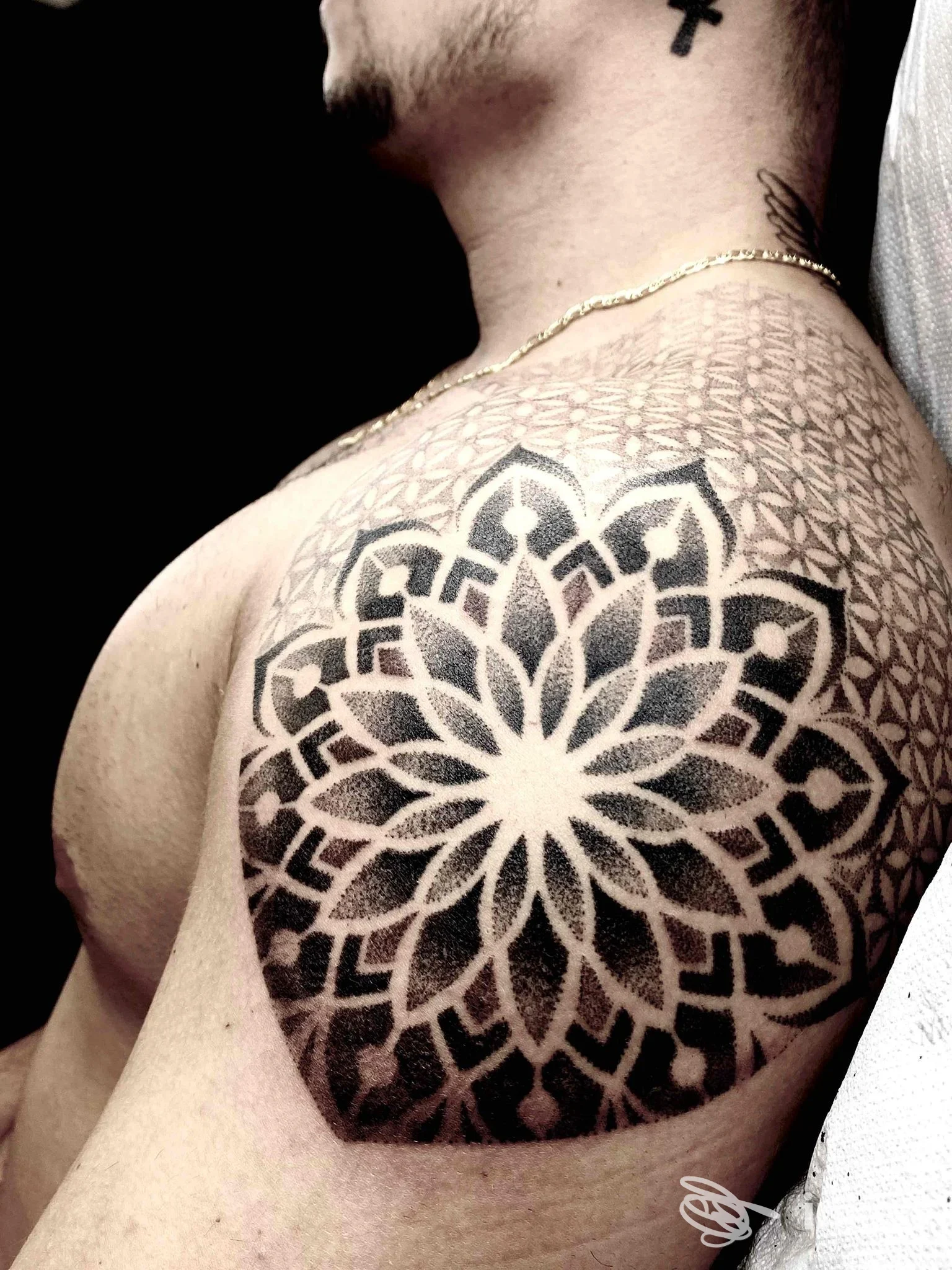 Parte superiore del braccio di un uomo, tatuaggio geometrico a mandala sulla spalla e parte della scapola, con pelle visibile e sfondo scuro.