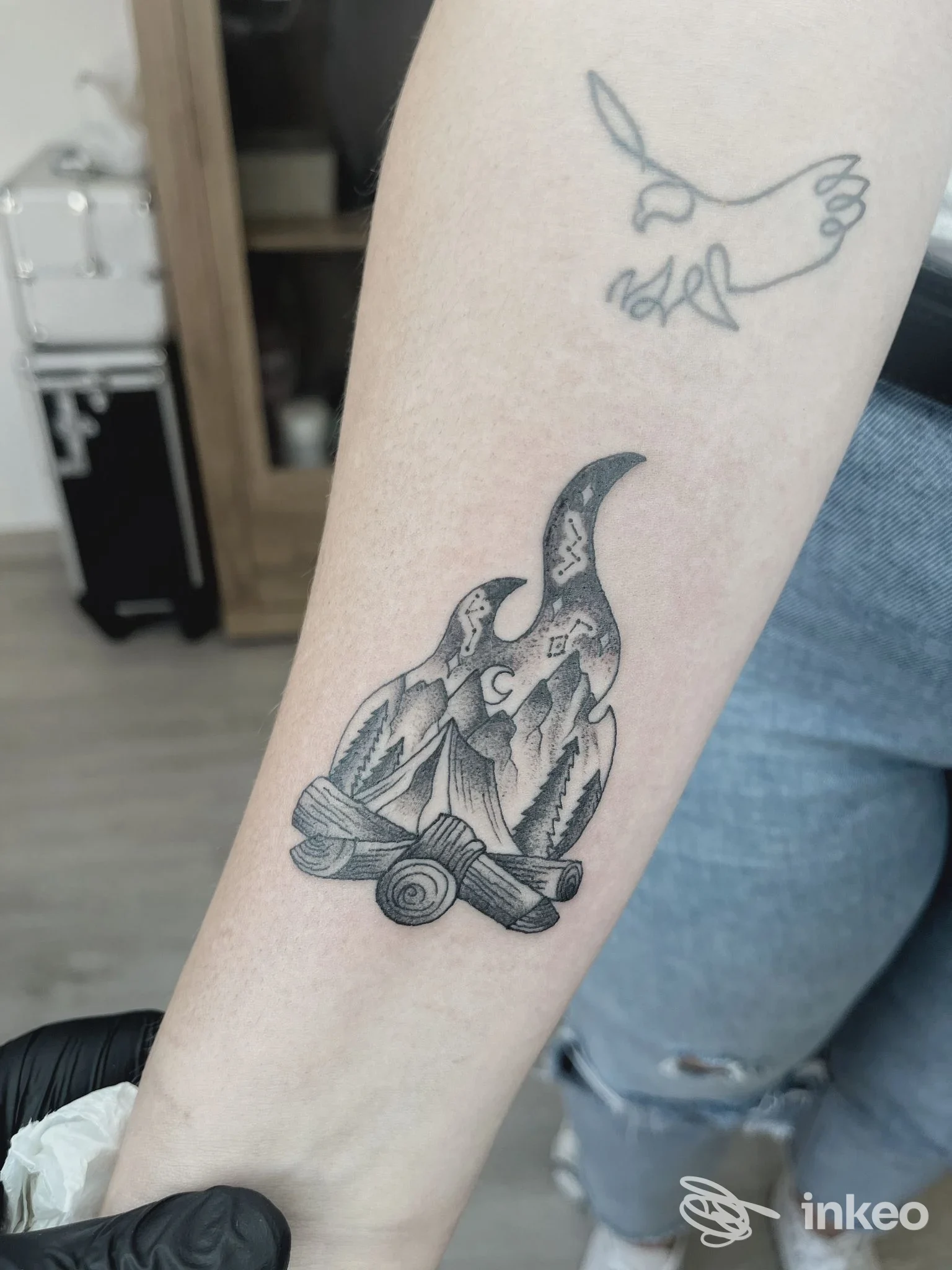 Avambraccio con tatuaggio nero/grigio: una fiamma che contiene una scena di montagne e tronchi, tatuaggio corporeo ben visibile su pelle chiara.