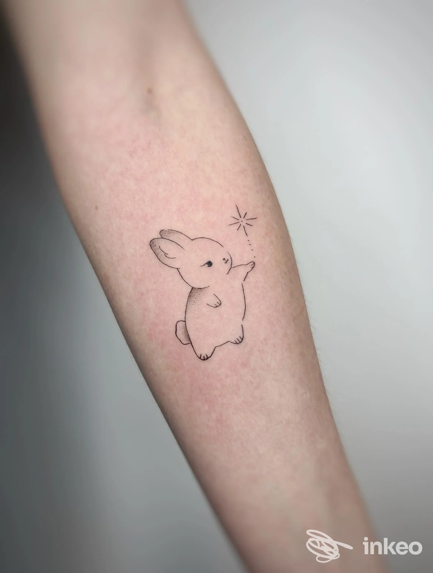 Tatuaggio lineare di un coniglietto che allunga una zampa verso una piccola stella sul braccio.