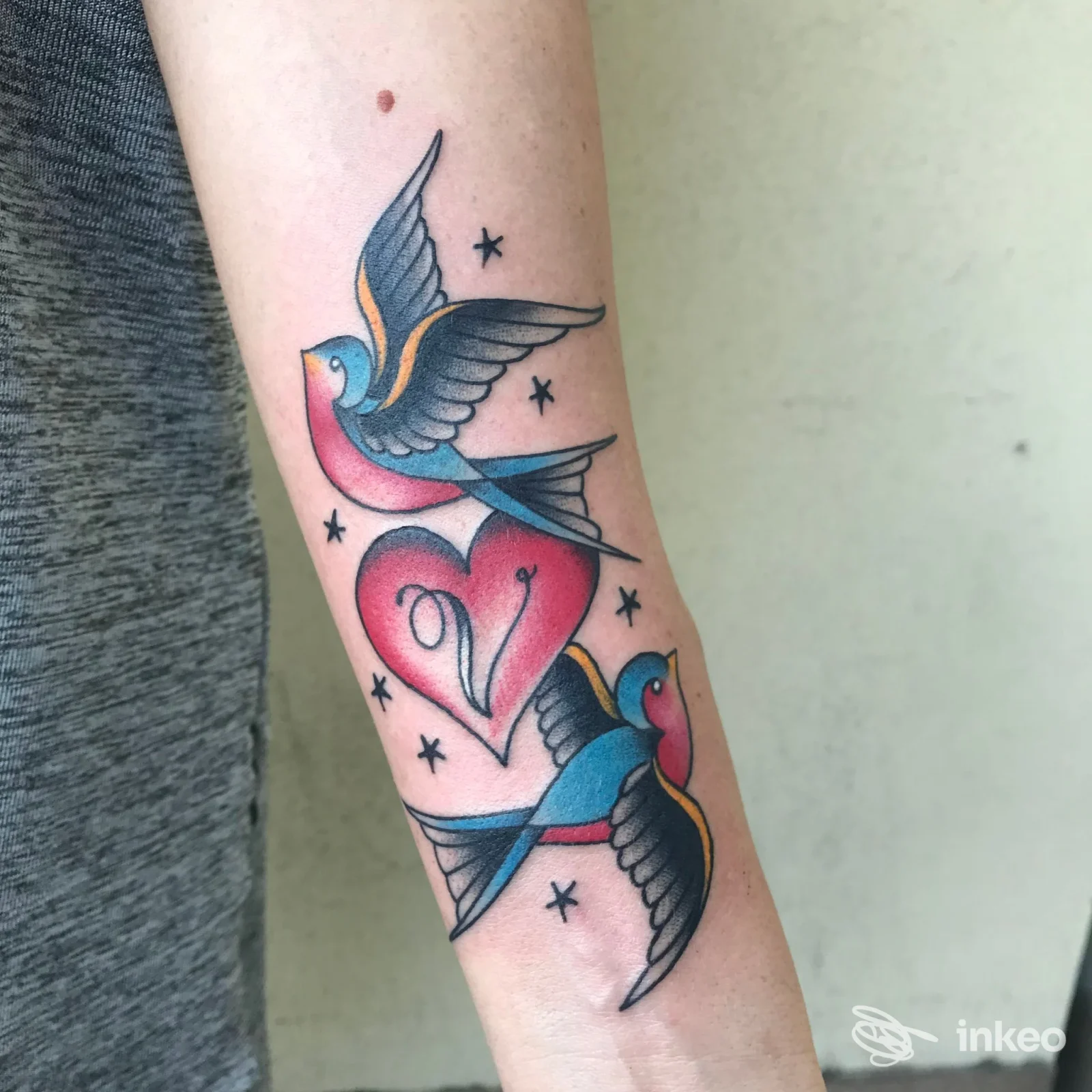Tattoo sul braccio realizzato da Ink Master Tattoo a Montesilvano: due colombe colorate intorno a un cuore, stelline nere e linee grafiche ben definite.