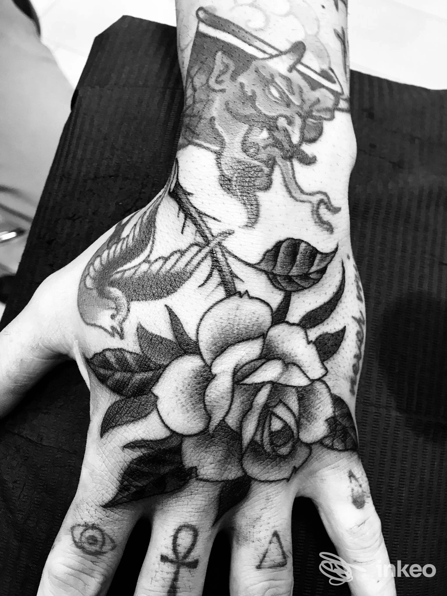 Mano sinistra con tatuaggio a fiore grande sul dorso, stile realistico in bianco e nero, visibile polso e dita, in un ambiente studio.