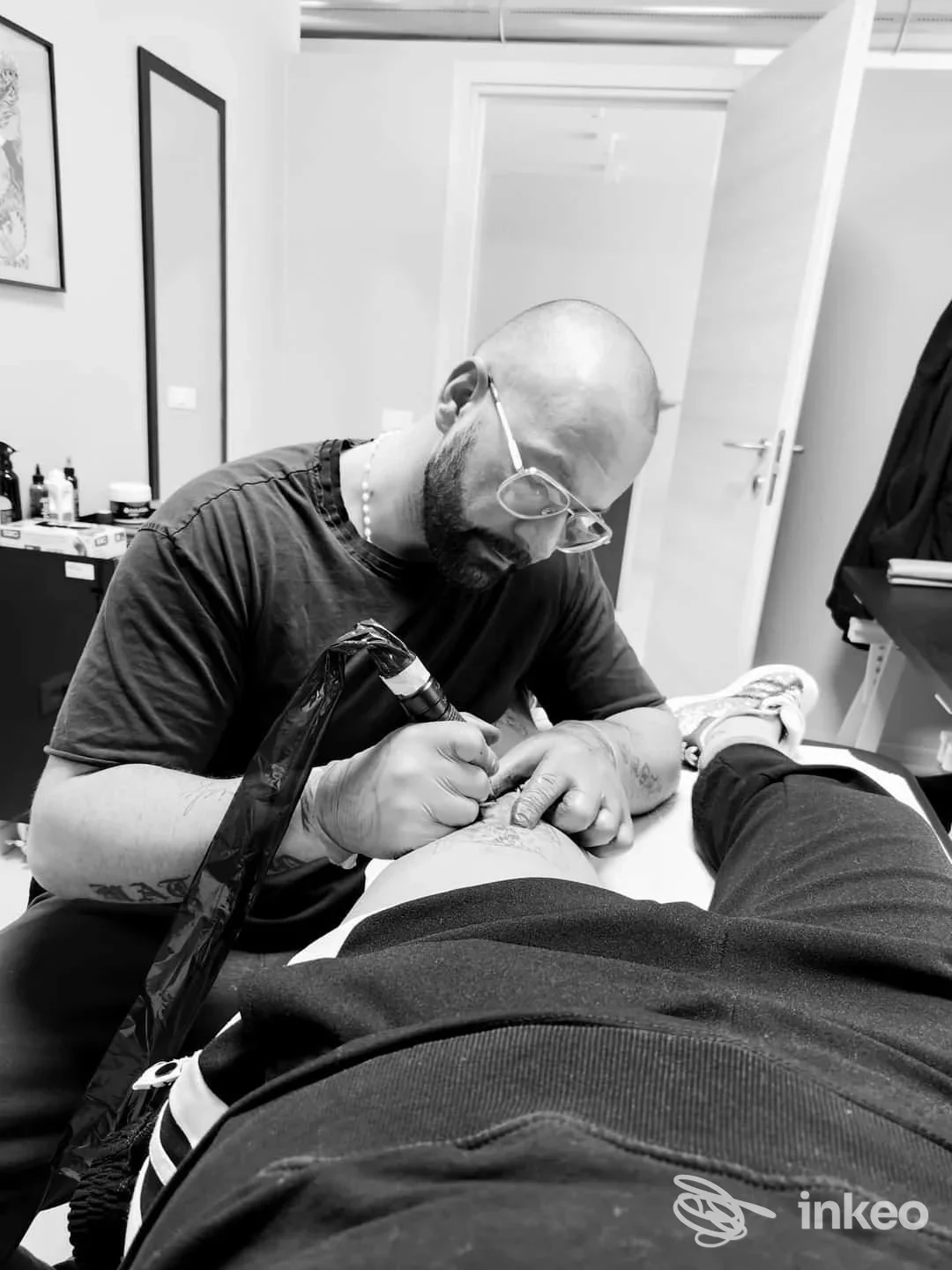 Un tatuatore con occhiali esegue un tatuaggio su una persona seduta, in un ambiente dello studio One Love Ink a Montesilvano, in bianco e nero