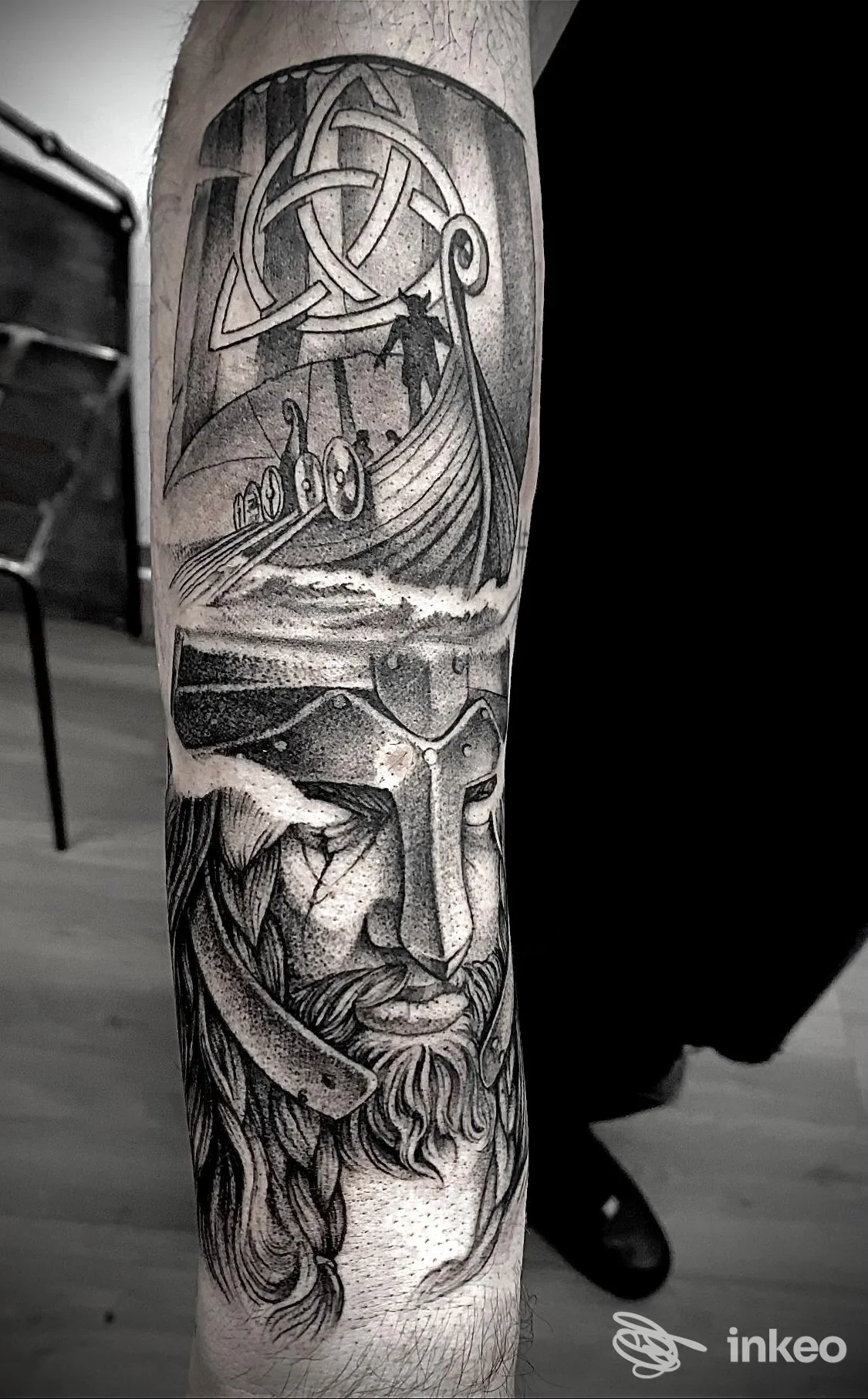 Tatuaggio realistico sull'avambraccio raffigurante un volto e un paesaggio marino con un motivo intrecciato celtico in alto; alto livello di dettaglio in bianco e nero su pelle.