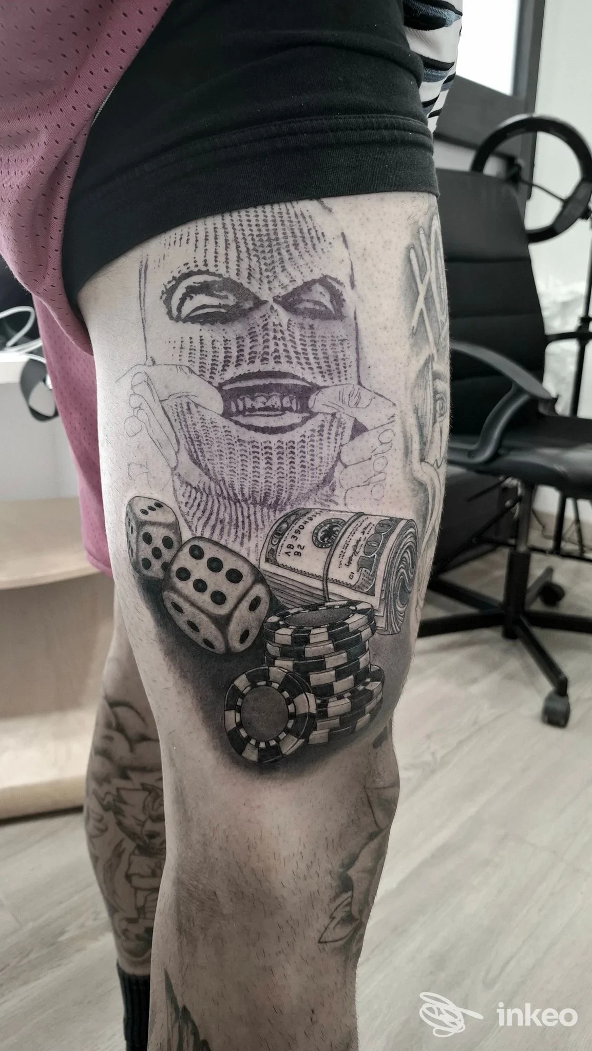 Tattoo visibile sulla coscia sinistra: faccia stilizzata con dadi e banconote, tatuaggio in bianco e nero su pelle umana.
