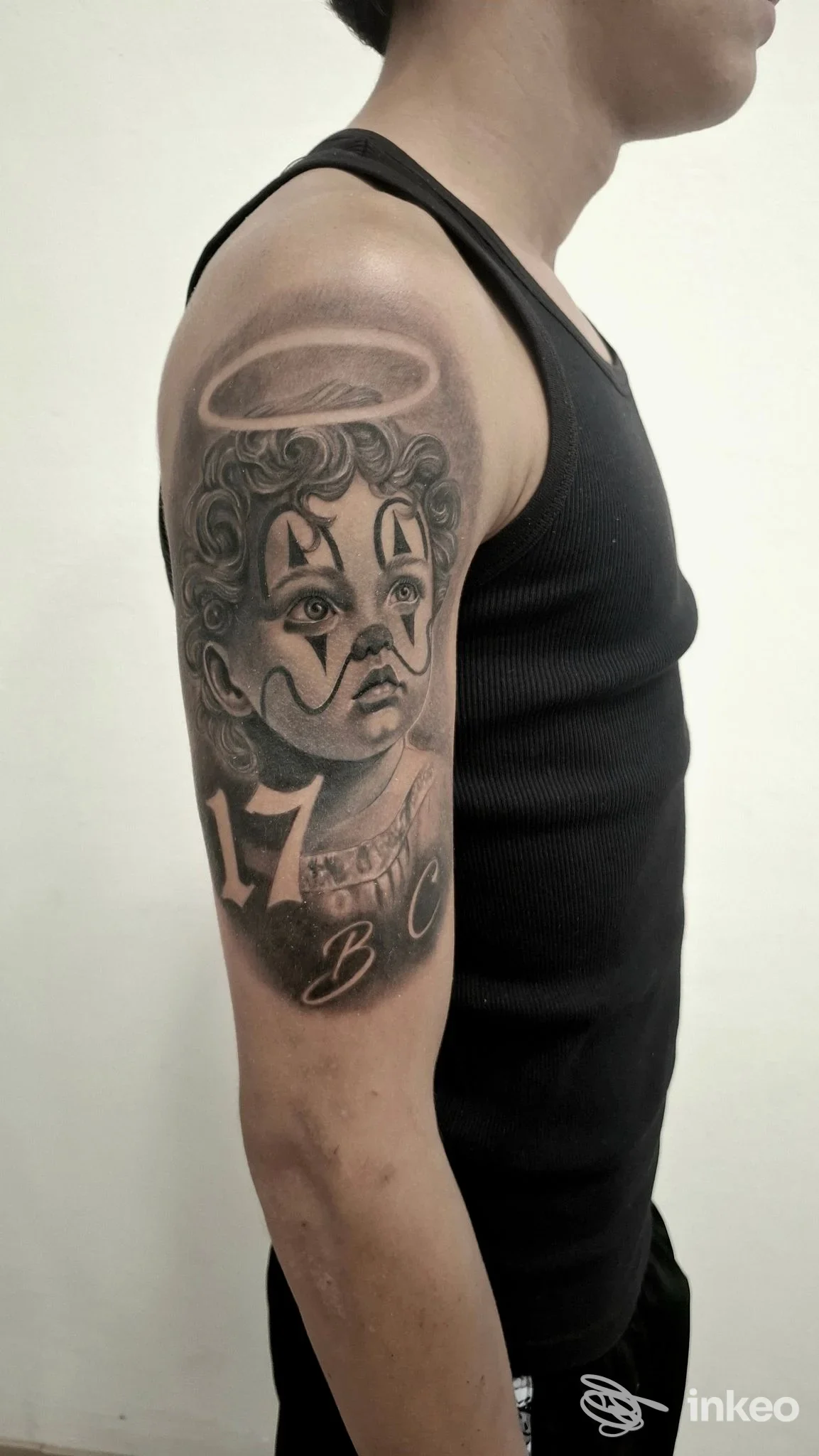 Tattoo sul braccio di una persona, ritratto stilizzato con aureola, numeri 17 e lettere BC; tatuaggio chiaramente visibile sulla pelle in una canotta nera.