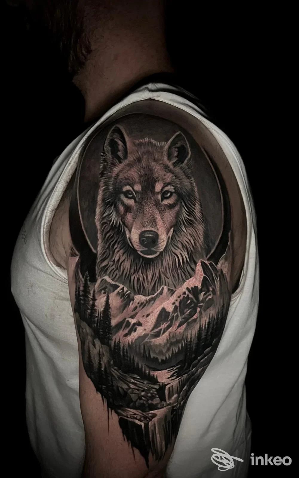 Tattoo realistico su spalla: volto di lupo all'interno di cerchio, con montagne sul braccio, in bianco e nero, scatto in controluce.
