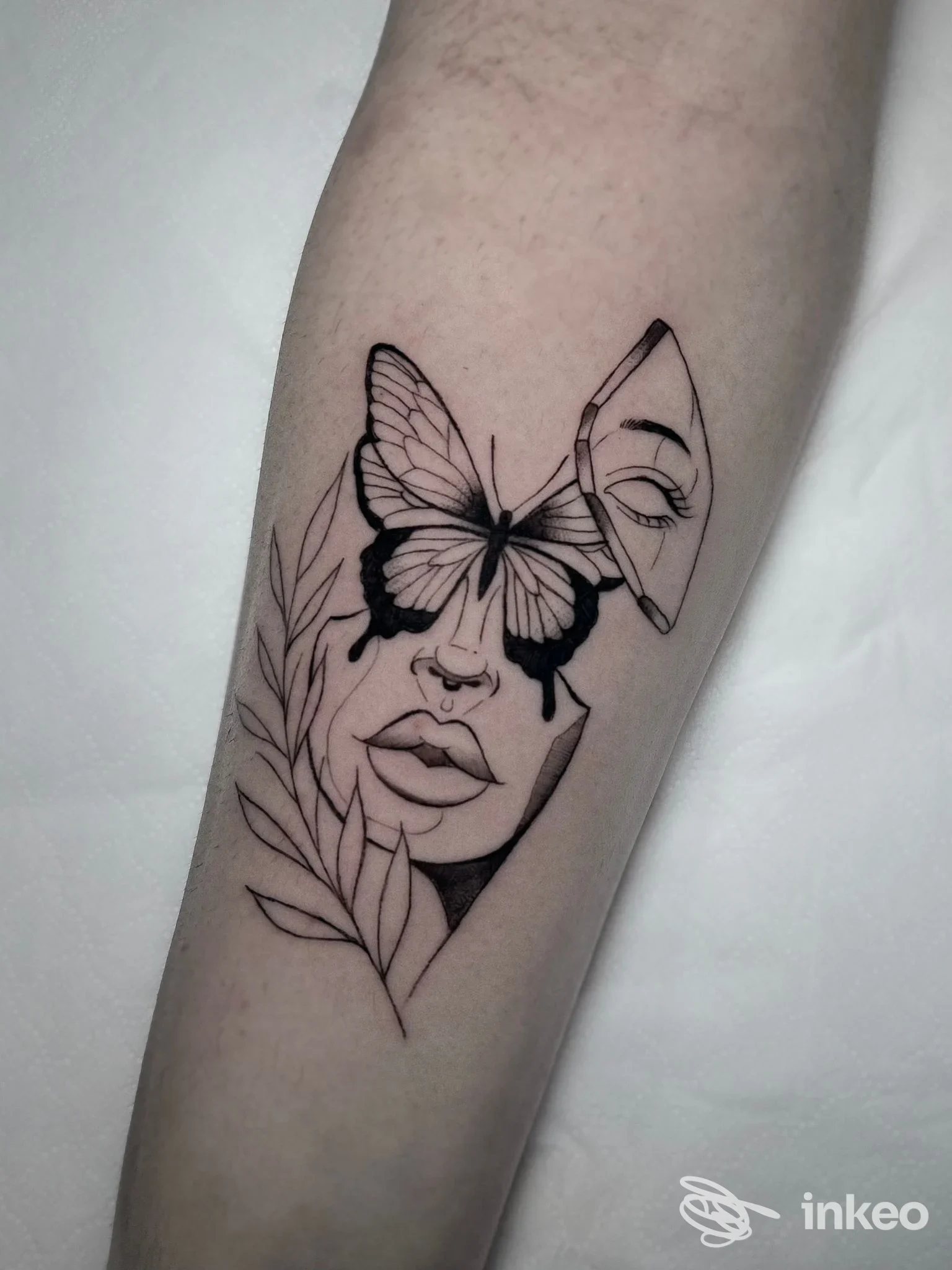 Tattoo su avambraccio raffigurante una figura femminile stilizzata con una farfalla che copre l'occhio, dettagli di foglie sul lato sinistro, eseguito con linee sottili e tratto nero.