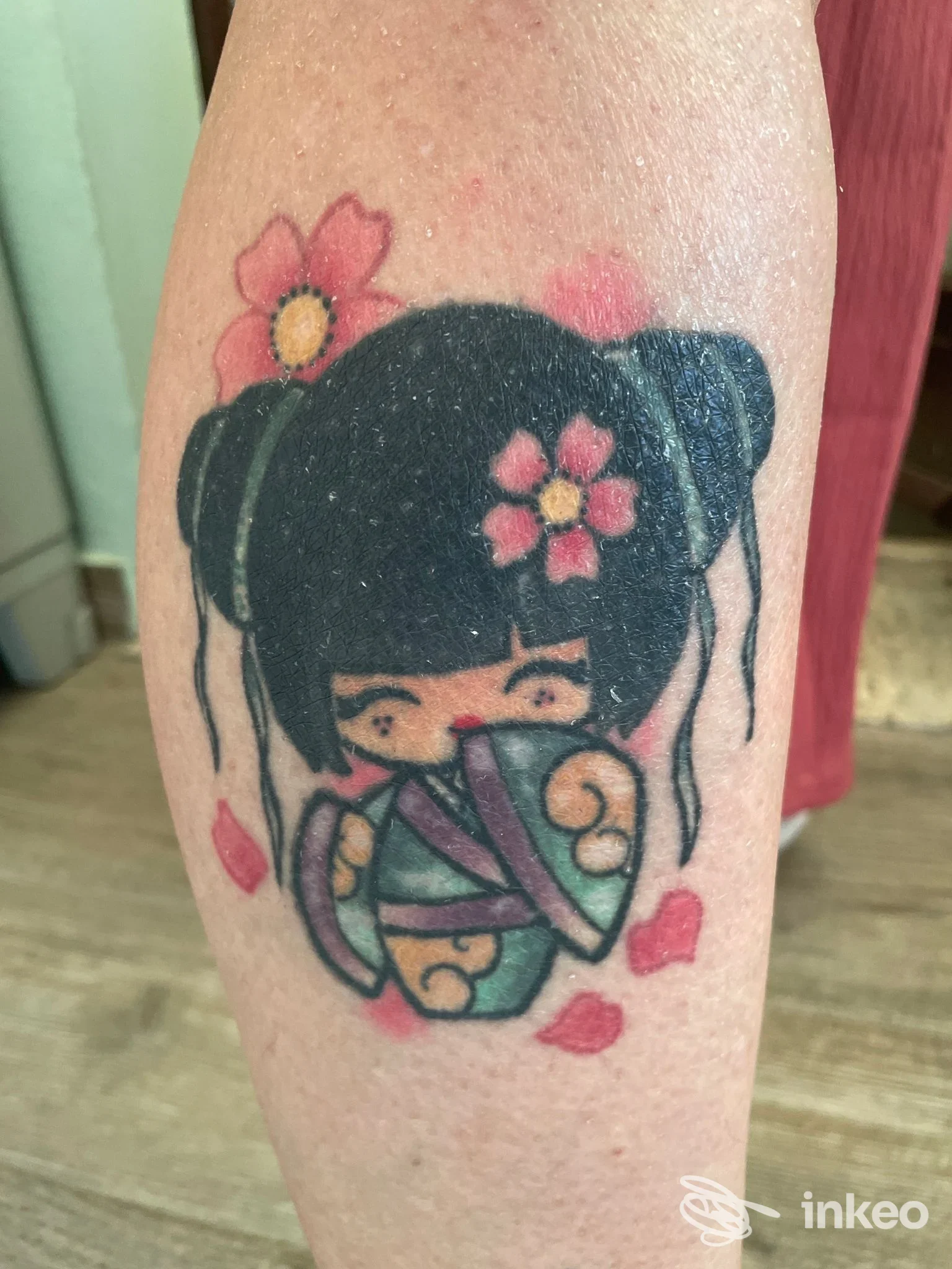 Logo di tatuaggio su avambraccio raffigurante una bambola giapponese con capelli neri, fiorellini rosa e kimono colorato, contorni netti e sfondo chiaro.