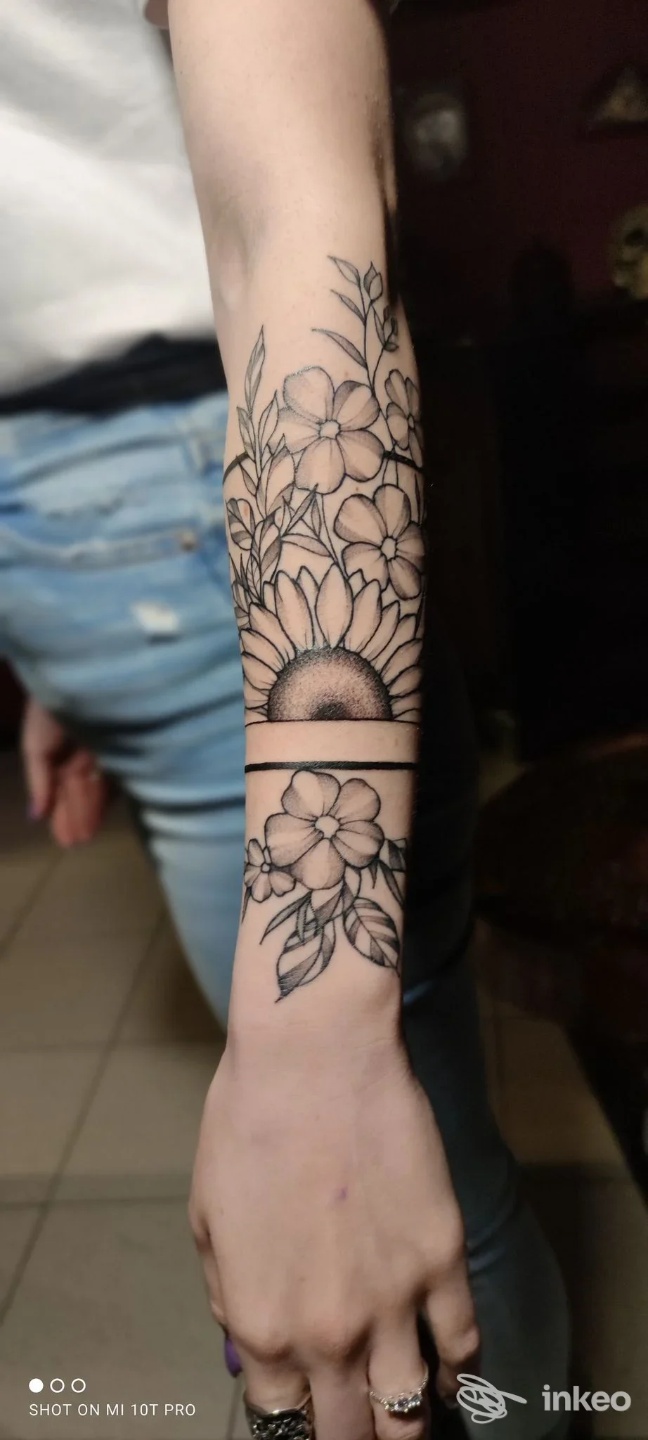 Tatuaggio floreale in bianco e nero sull'avambraccio sinistro, con bouquet di fiori e una semicirconferenza centrale.