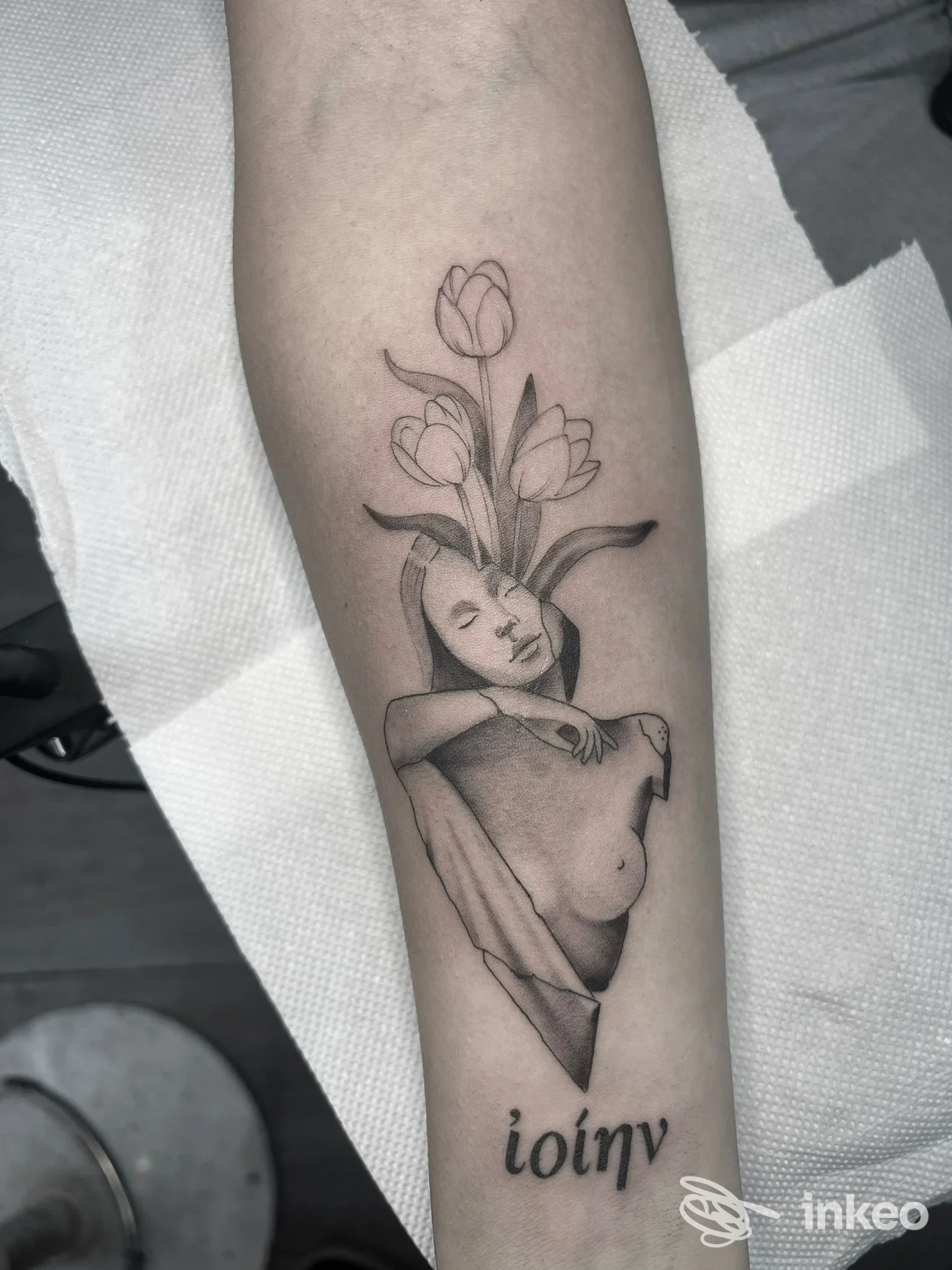 Tattoo su avambraccio raffigurante figura femminile in stile disegnato, con tulipani che emergono dalla testa; tonalità nere e grigie, senza testo o elementi di overlay.