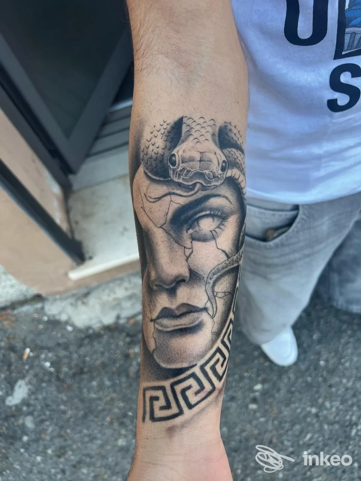 Tacita foto di un braccio con tatuaggio realistico che mostra un volto e elementi ornamentali, con motivi geometrici lungo l’avambraccio, tonalità in bianco e nero.
