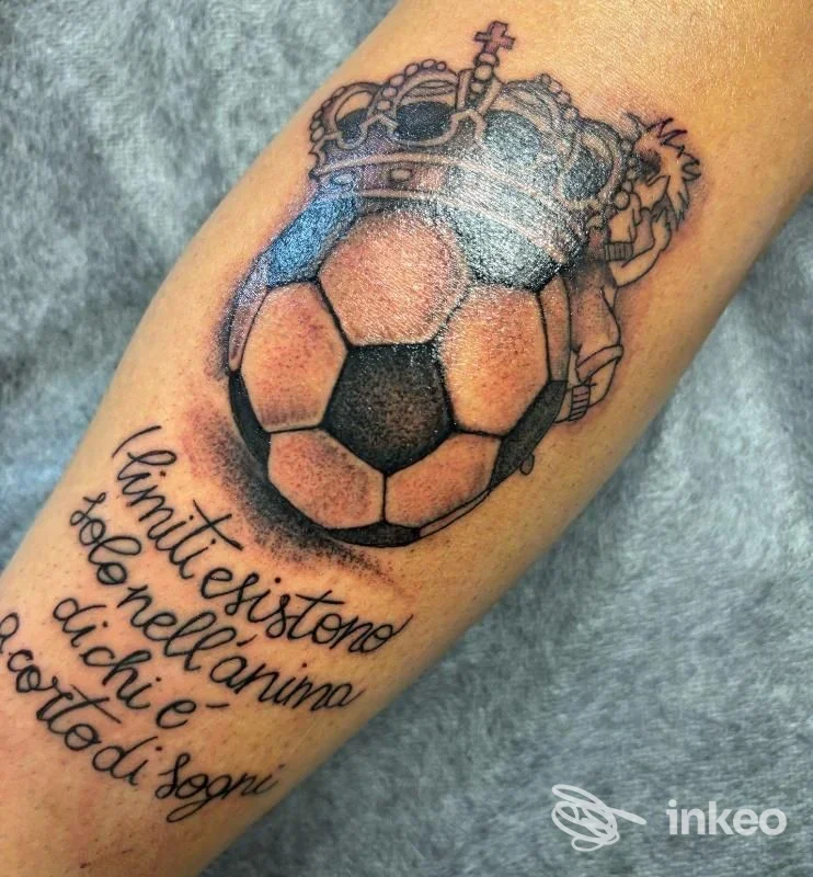 Tattoo su avambraccio raffigurante una palla da calcio con corona sopra, accompagnata da testo chiamato in corsivo. Il disegno è in bianco e nero con linee nette.
