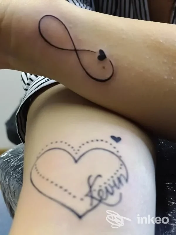 Due tatuaggi visibili sulla pelle: un motivo infinito con piccolo cuore sul braccio e un grande cuore con bordi tratteggiati sul polpaccio, entrambi in stile lineare nero.