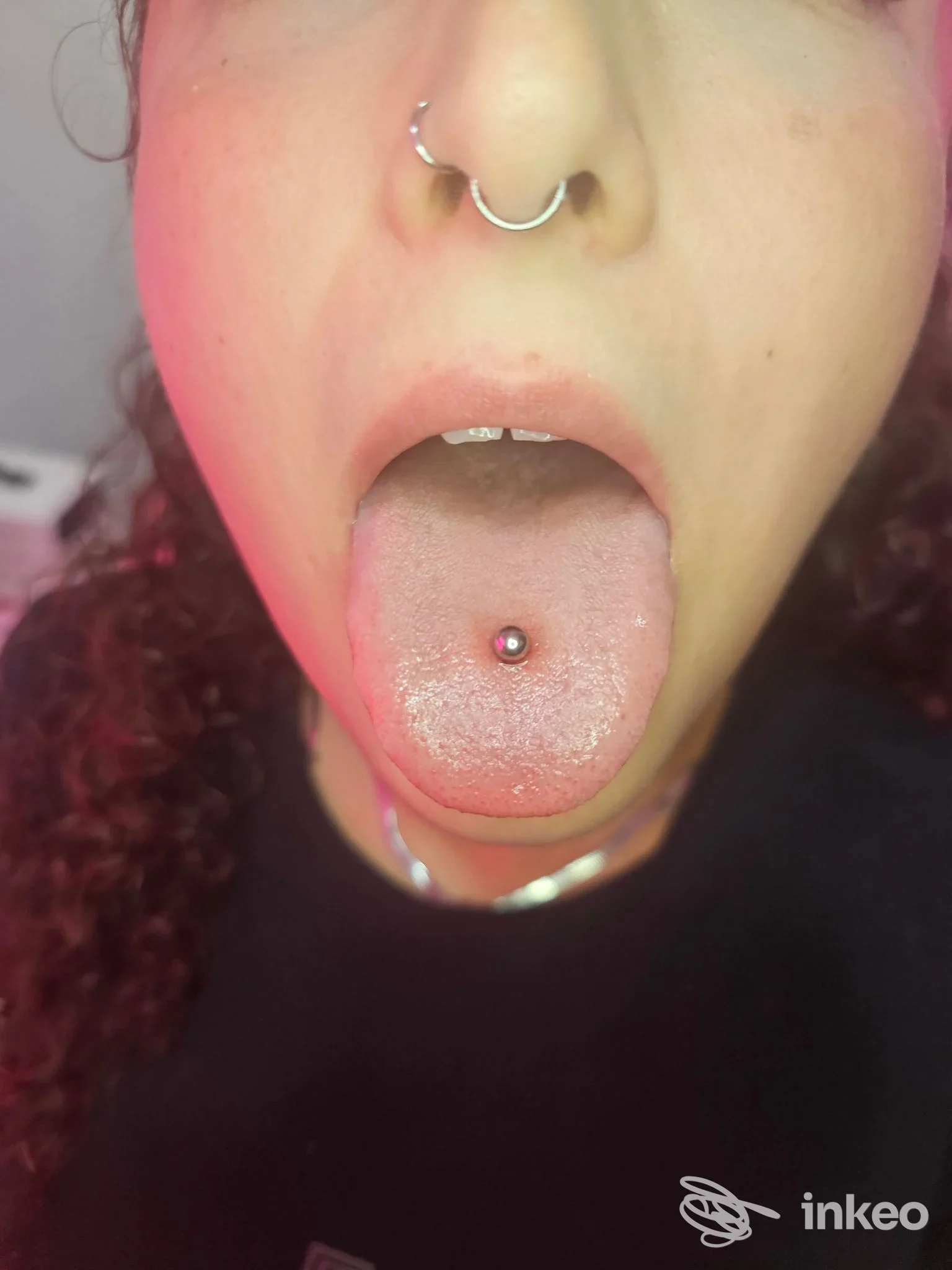 Piercing alla lingua ben visibile in primo piano, anello a perno dorato attraverso la lingua, sullo sfondo si intravede una persona; fotografia promossa dallo studio Baruna Piercer a Reggio di Calabria.