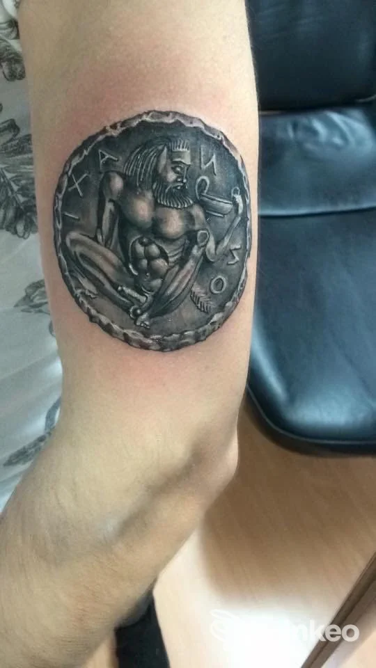 Tattoo circolare sul braccio raffigurante un guerriero muscoloso con torace nudo, in tonalità nere e grigie, cornice decorativa.