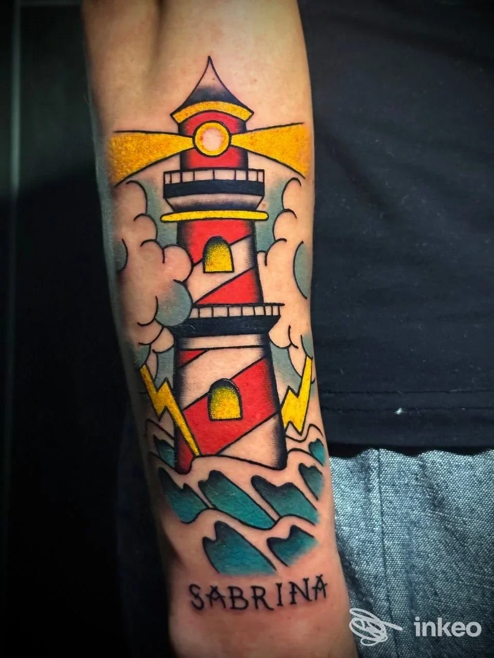 Tattoo su braccio raffigurante un faro colorato con onde azzurre, firma Sabrina in basso; eseguito dallo studio Kalabria Ink a Botricello.