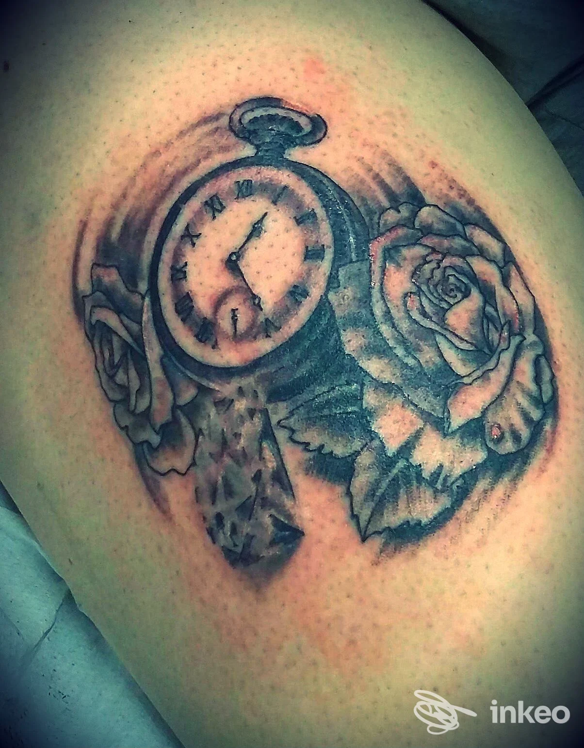 Tatuaggio realistico in bianco e nero raffigurante una bussola-orologio circondata da rose, applicato sulla pelle della spalla.