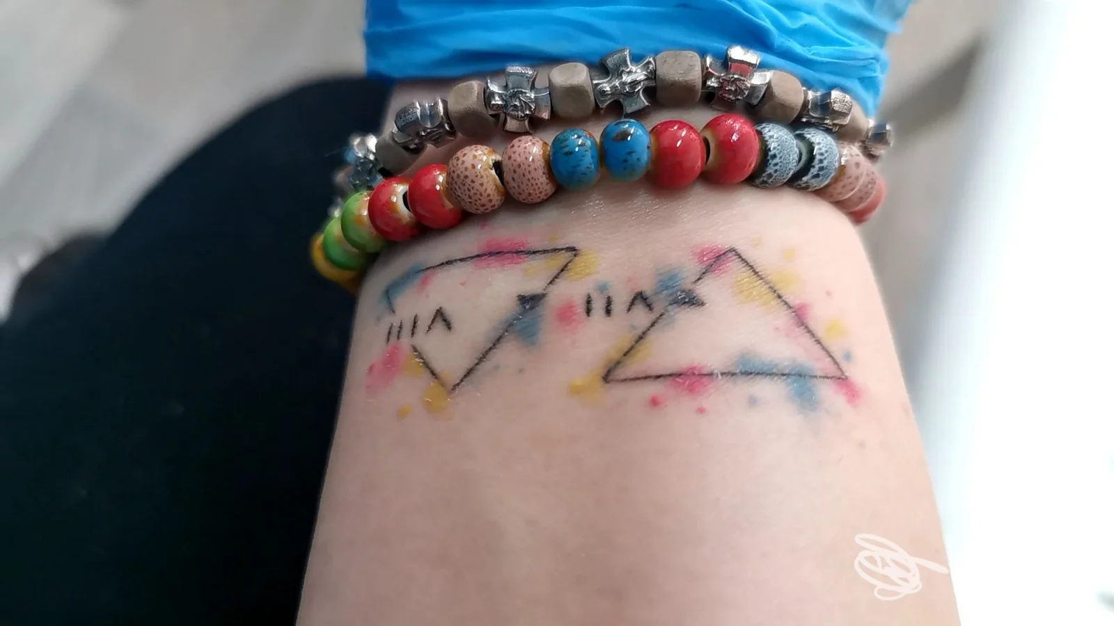 Due triangoli geometrici colorati disegnati sulla pelle della persona, con contorni neri e riflessi rosa, visibili sull'avambraccio o sulla gamba; braccialetti colorati sopra la pelle.