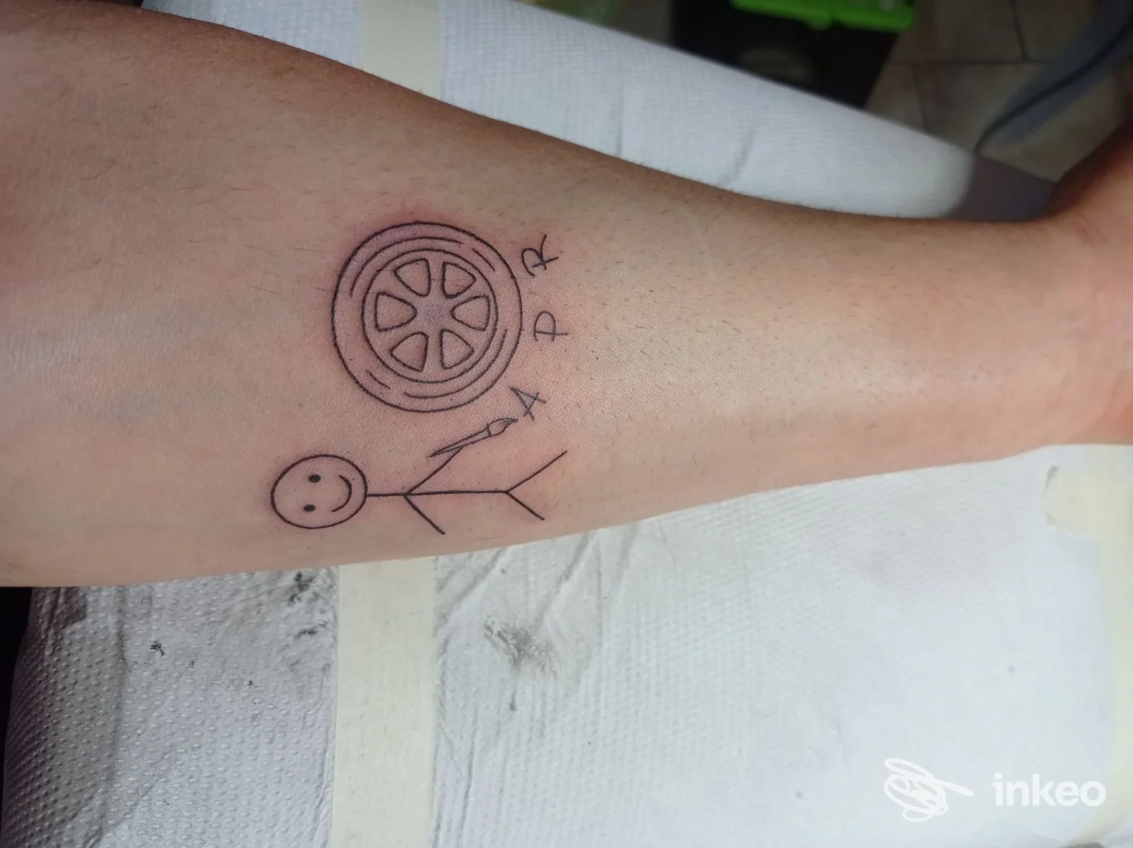 Tatuaggio a linea sull'avambraccio: figura stilizzata con testa a cerchio e corpo a linee, accanto a una ruota con cerchi. visibile sul braccio destro, in uno studio di tatuaggi a Girifalco.