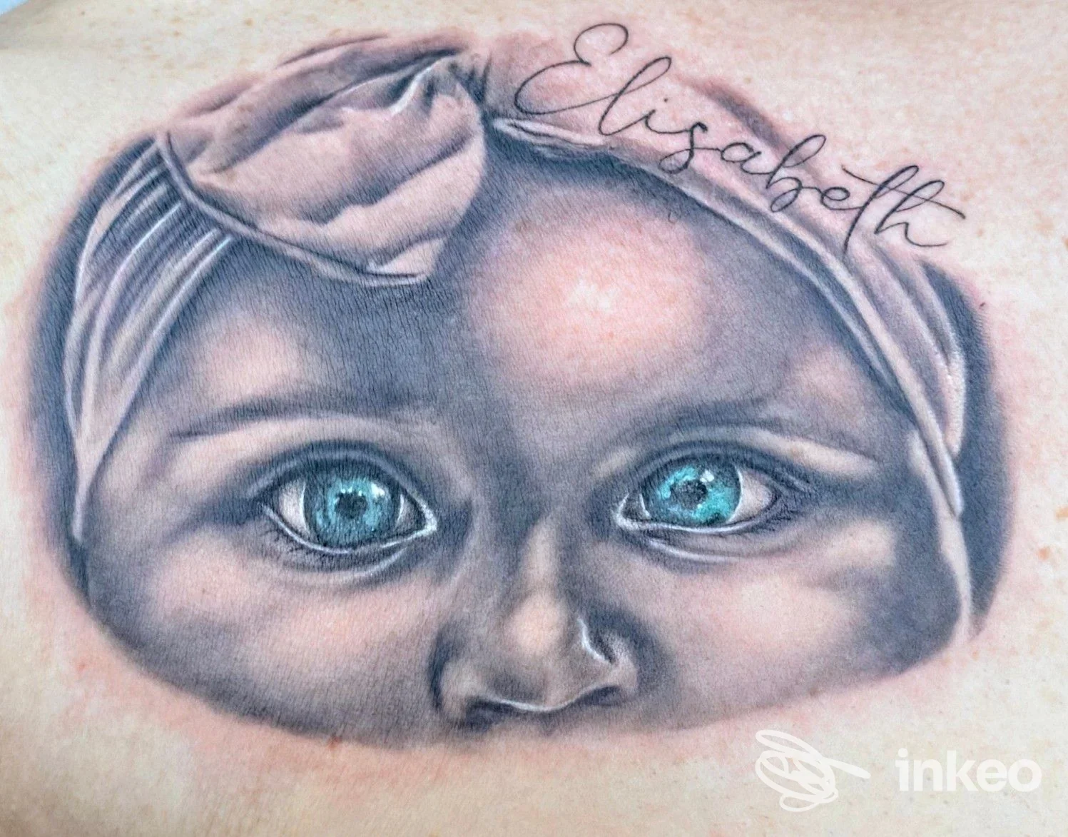 Ritratto realistico di un volto di bambino tatuato sulla pelle, con occhi azzurri e una fascia in testa; firma in corsivo sopra la fronte, visibile sullo stesso pezzo di pelle.