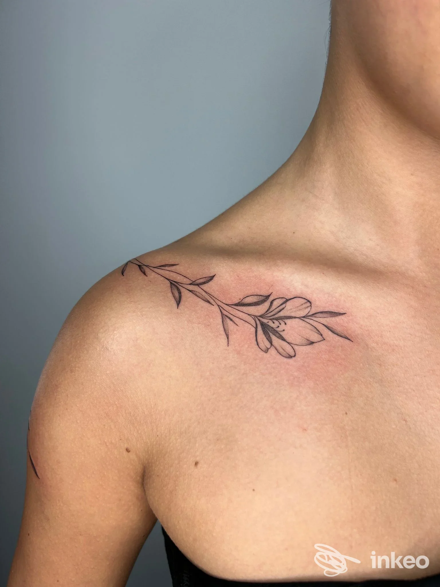 Tattoo floreale lineare lungo la clavicola e la spalla, in nero, su pelle chiara, parte di un braccio/torace visibile.