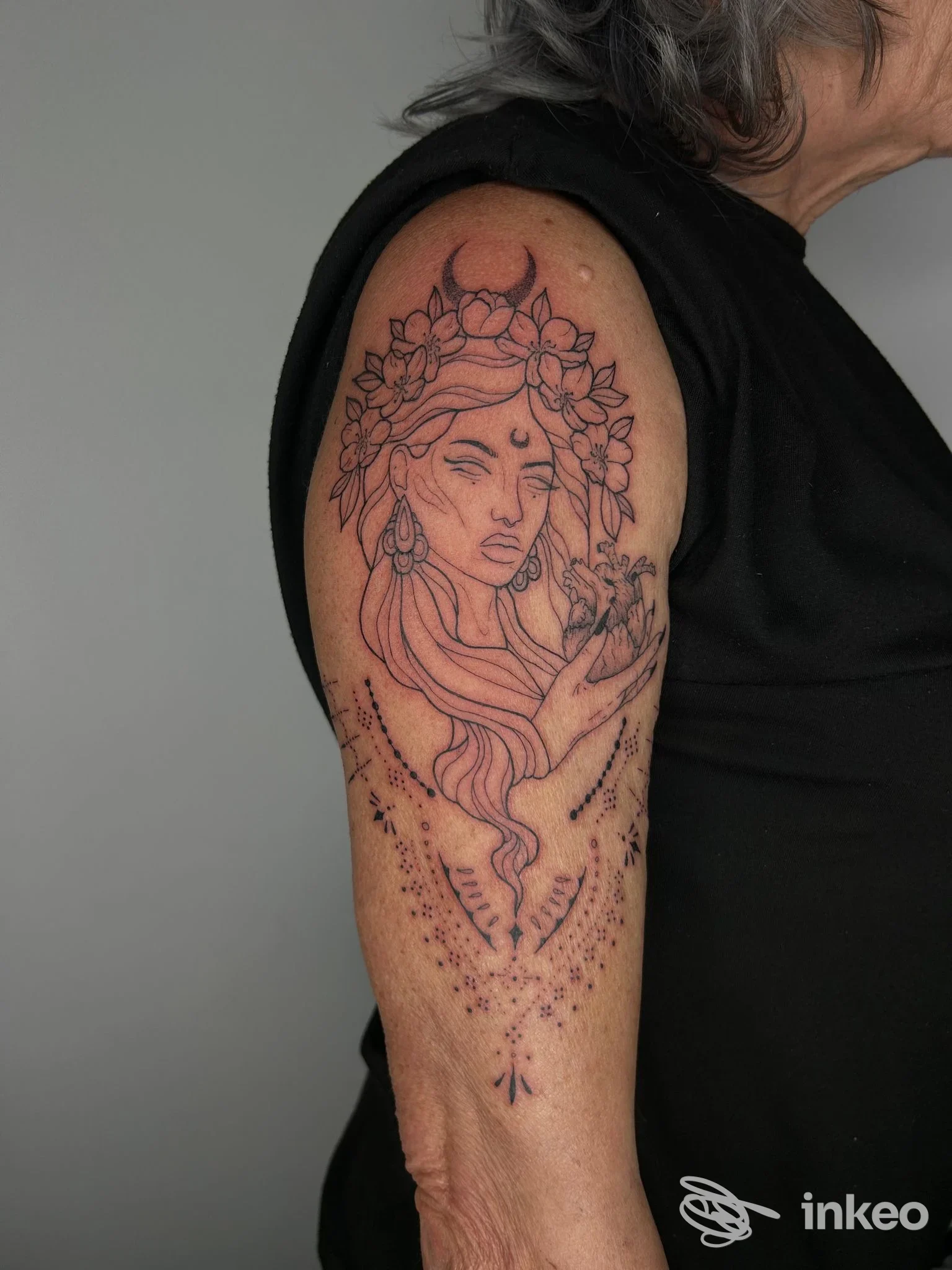 Tattoo a tema femminile sul braccio visibile: linea nera, corpo e fiori intorno alla figura, viso in profilo parziale e strisce decorative sotto il braccio.