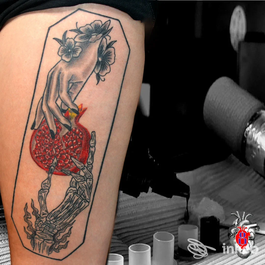 Tatuaggio colorato raffigurante una mano che sostiene un melograno, fiori e rami, inciso su coscia, incorniciato da bordi a forma di sarcofago.