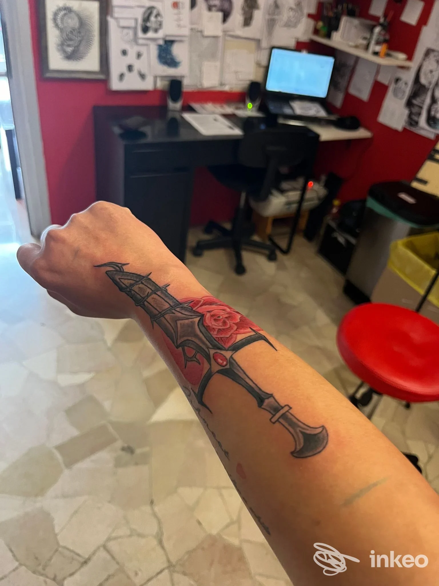 Tatuaggio sull’avambraccio raffigurante una lama nera con riflessi accanto a una rosa rossa, visibile su pelle reale.