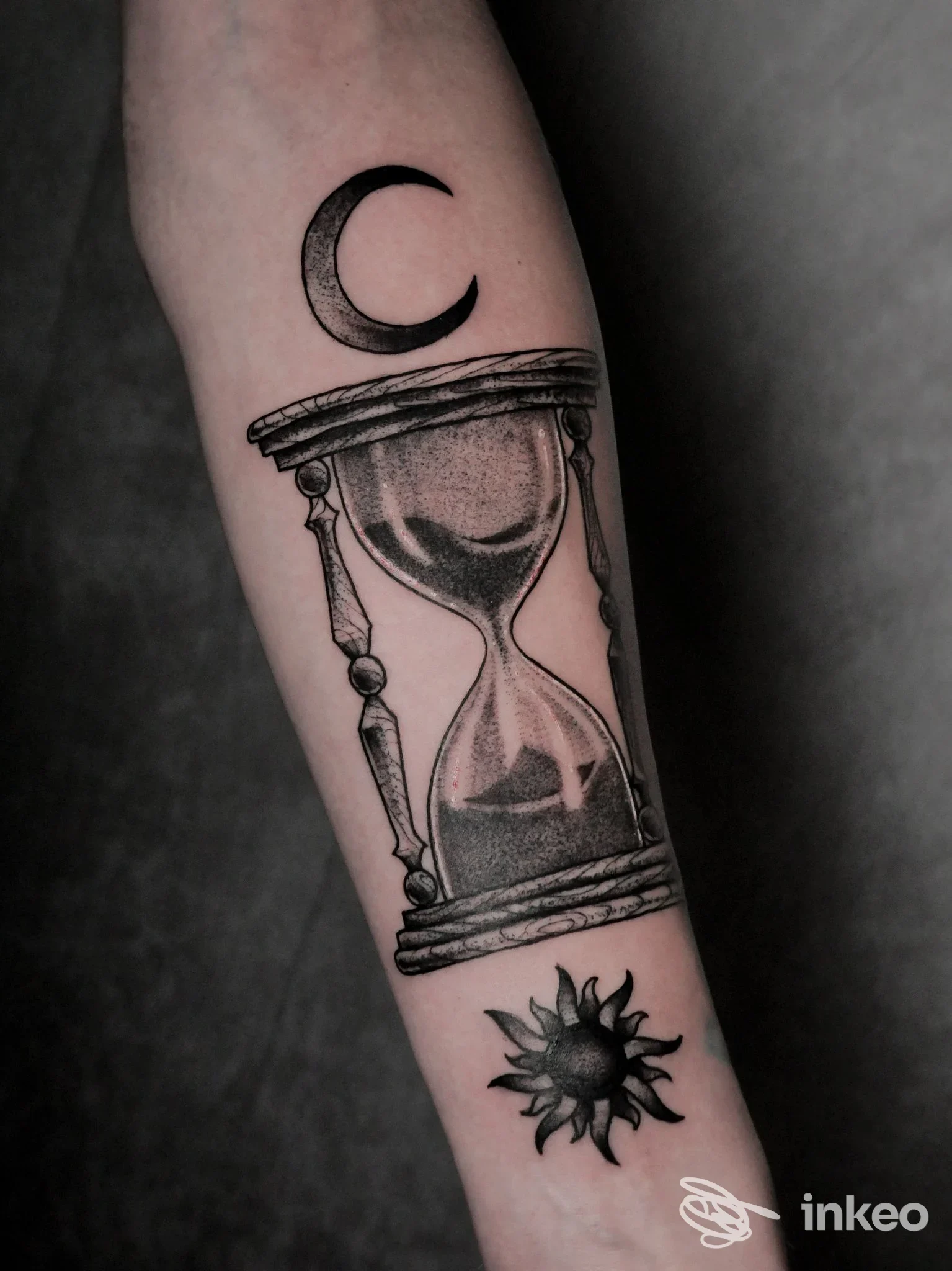 Tattoo sull'avambraccio raffigurante una clessidra con dettagli in ombreggiatura, una luna crescente sopra e un sole stilizzato sotto, realizzato con linee nette e contrasti neri.