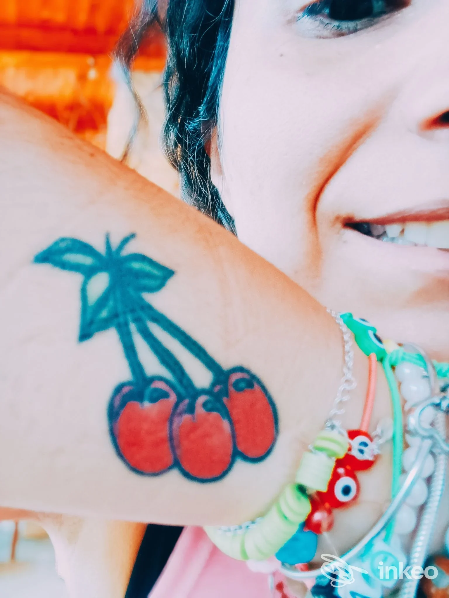 Braccio di una persona con tatuaggio di una coppia di ciliegie rosse e una palma stilizzata in blu, accompagnato da braccialetti colorati.