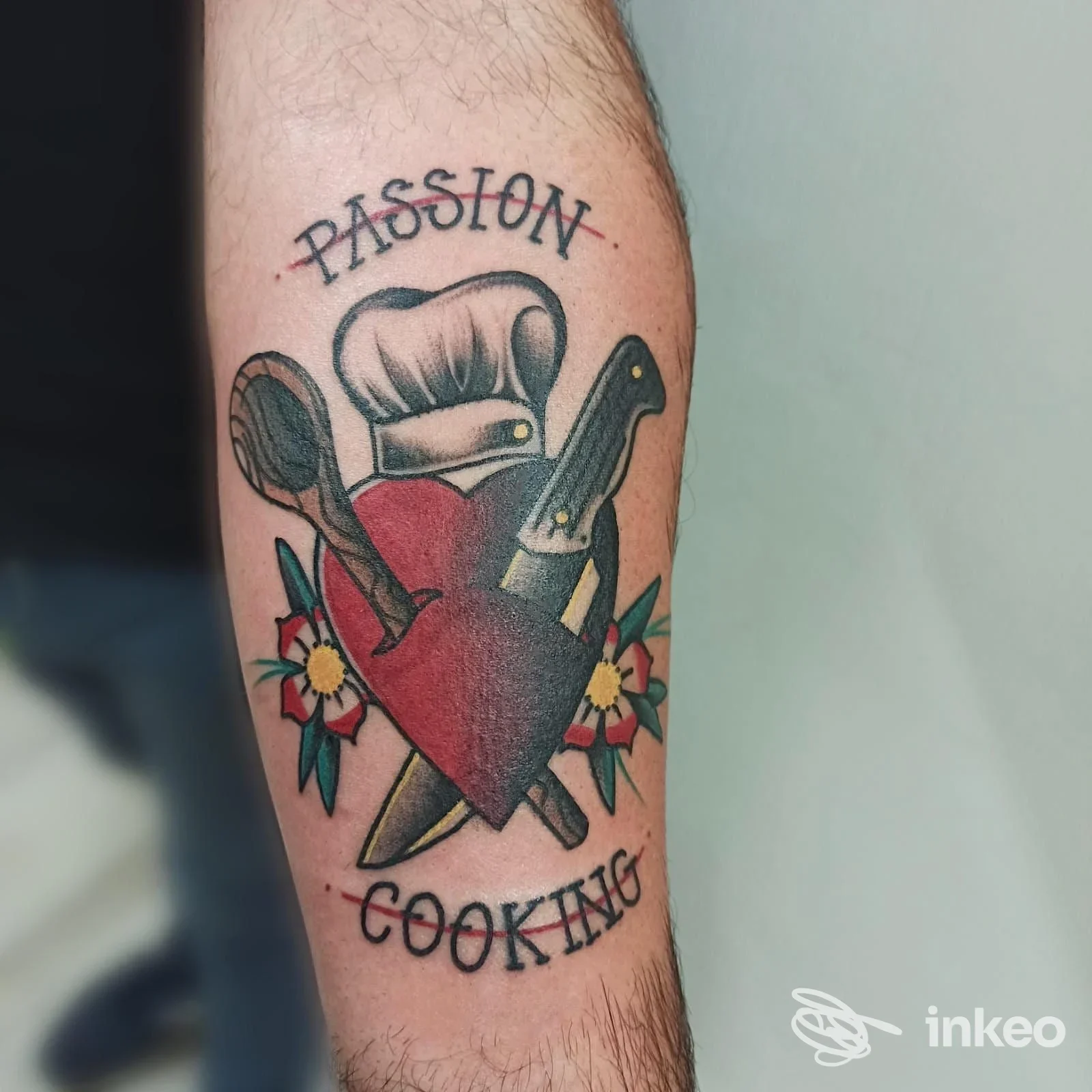 Tatuaggio sul braccio raffigurante cuore rosso con cappello da chef, forbice e coltello incrociati, accompagnati da fiori, tono colori vividi.