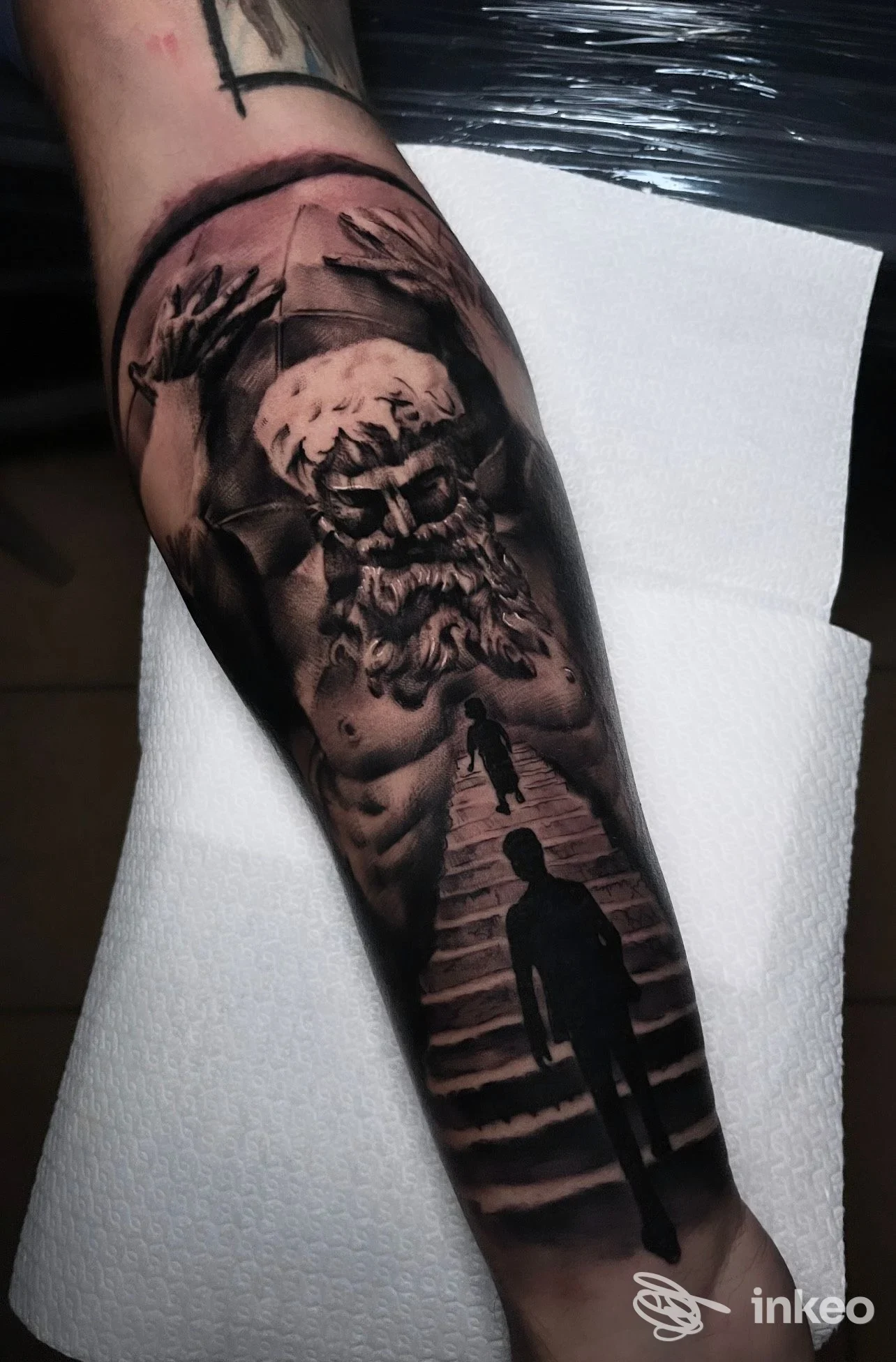 Tatuaggio in bianco e nero sull'avambraccio, raffigurante figure su uno sfondo scuro, realizzazione corporeale nitida e ben definita.