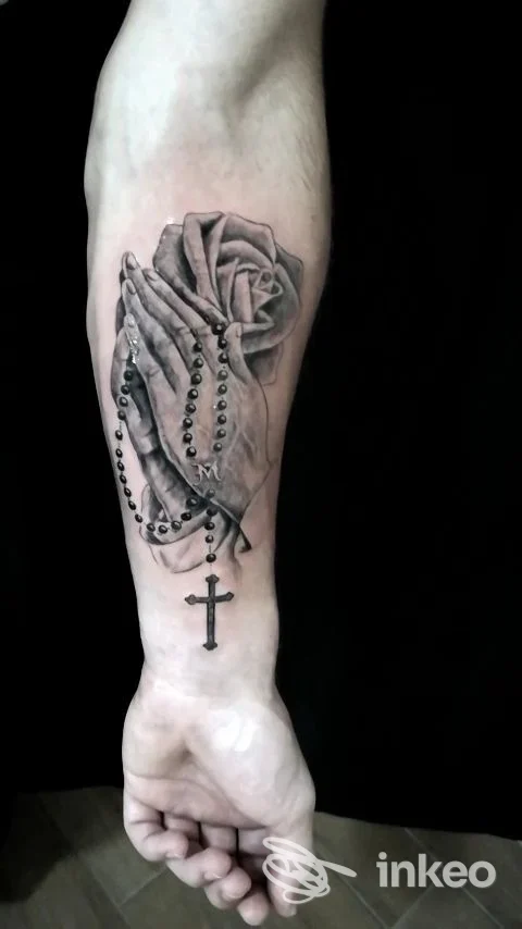 Tattoo sull'avambraccio raffigurante una rosa con rosario a catena e croce pendente, in tonalità scure su pelle chiara.