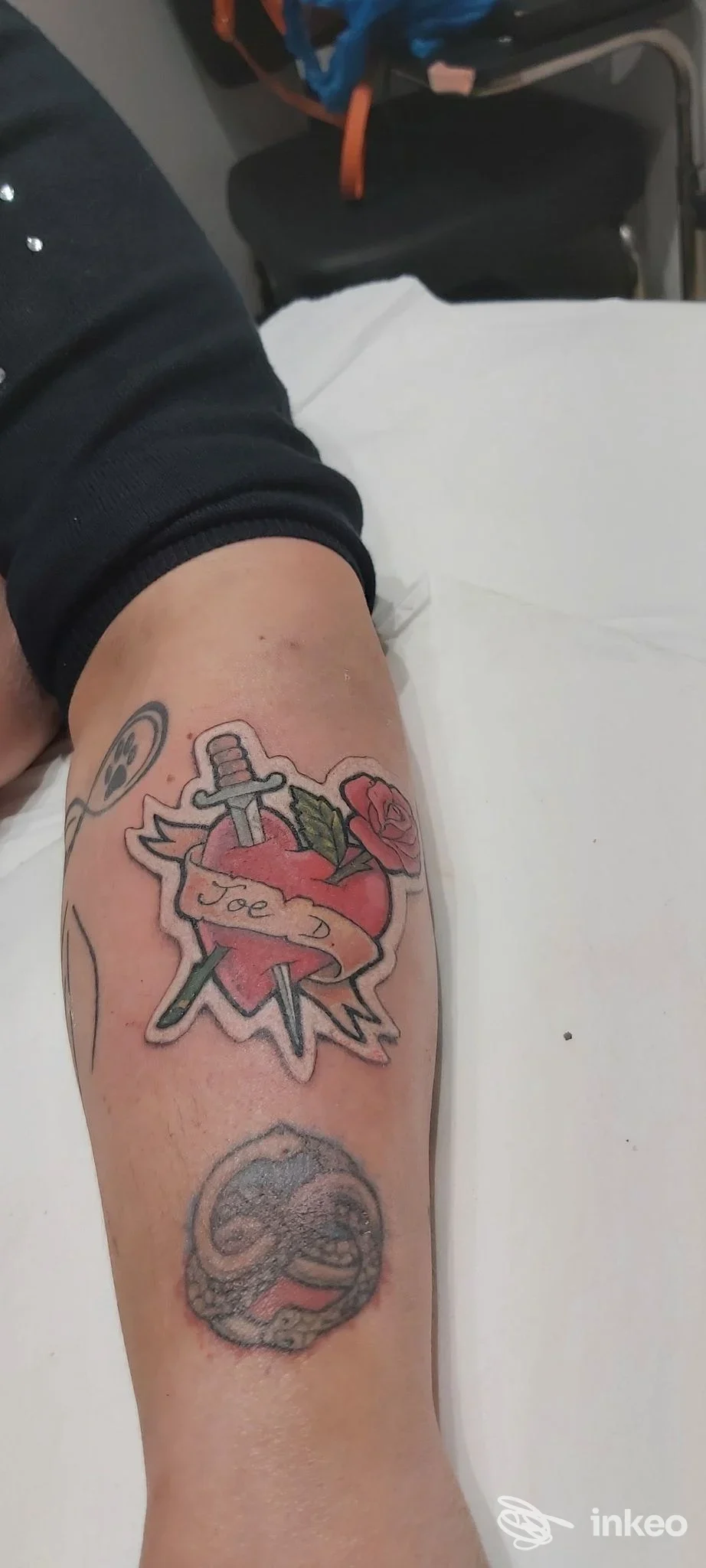 Tatuaggio colorato sull'avambraccio: cuore con lama e rose, con nastro e basse tonalità, senza volto visibile.