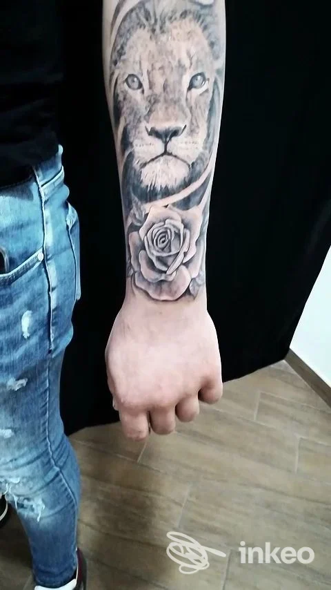 Tattoo realistico di un leone sul polso/avambraccio con una rosa sotto, realizzato sulla pelle di una persona. tonalità in bianco e nero/grigi, dettaglio definito.