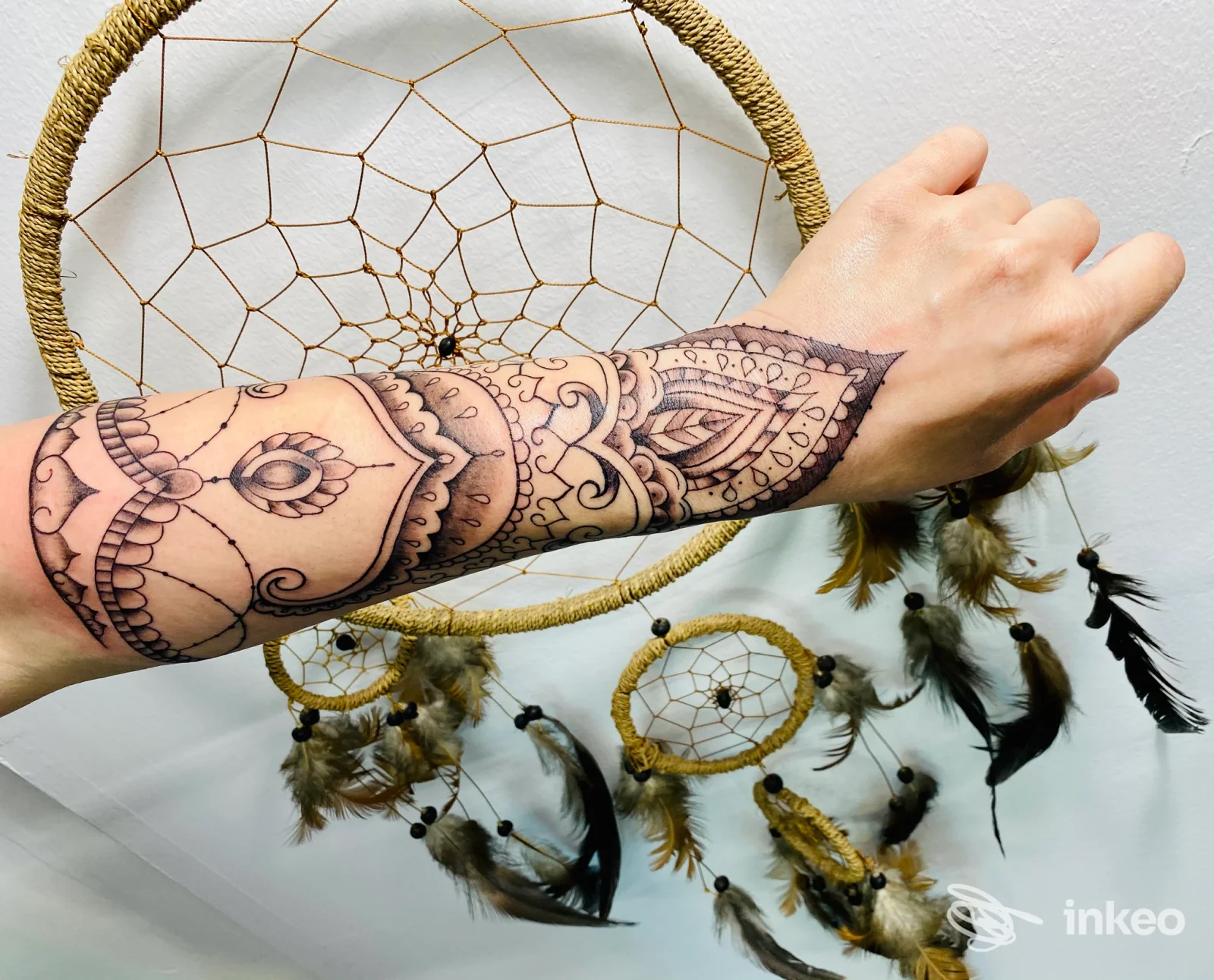 A sinistra, un avambraccio con un tattoo esteso che mostra motivi ornamentali e linee geometriche scure; sfondo sfumato con cerchi speziati appesi.