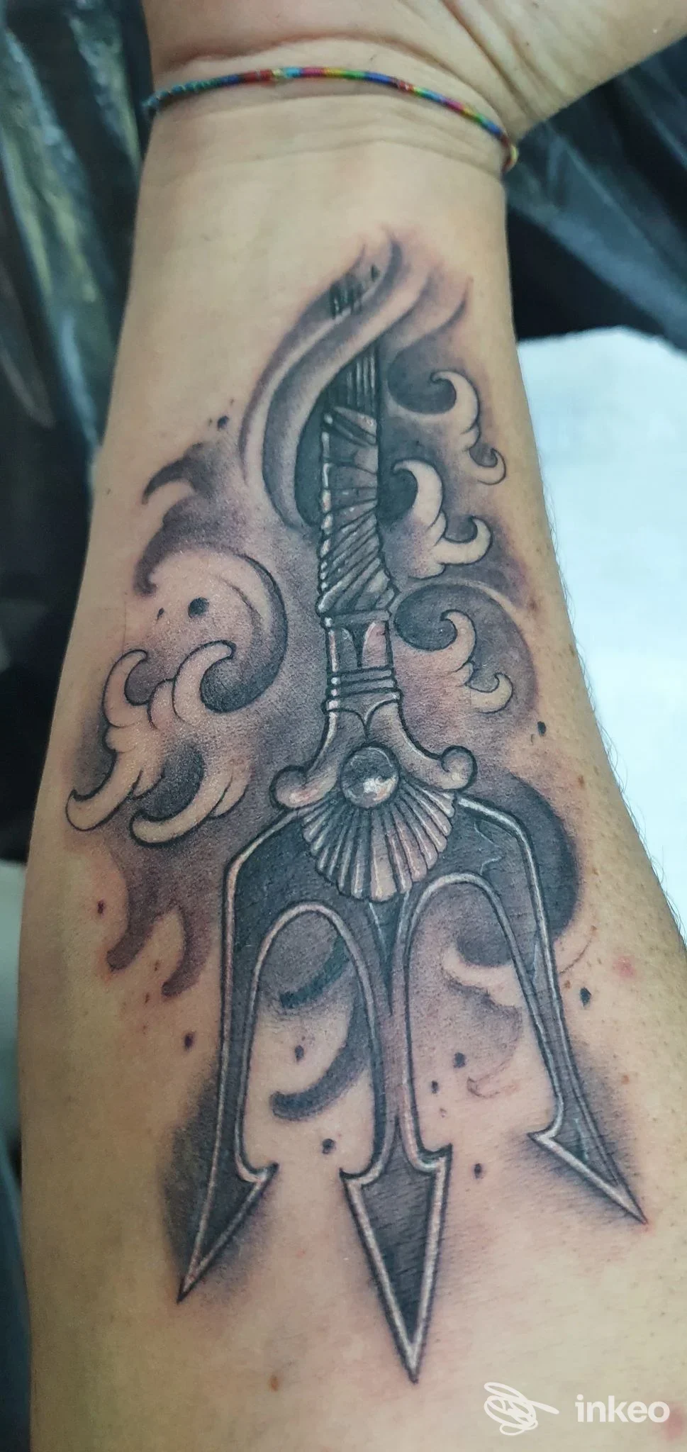 Tattoo visibile sull’avambraccio: una lama appuntita o tri-prong in stile grafico, con sfumature in grigio e contorni marcati, sfondo sfumato.