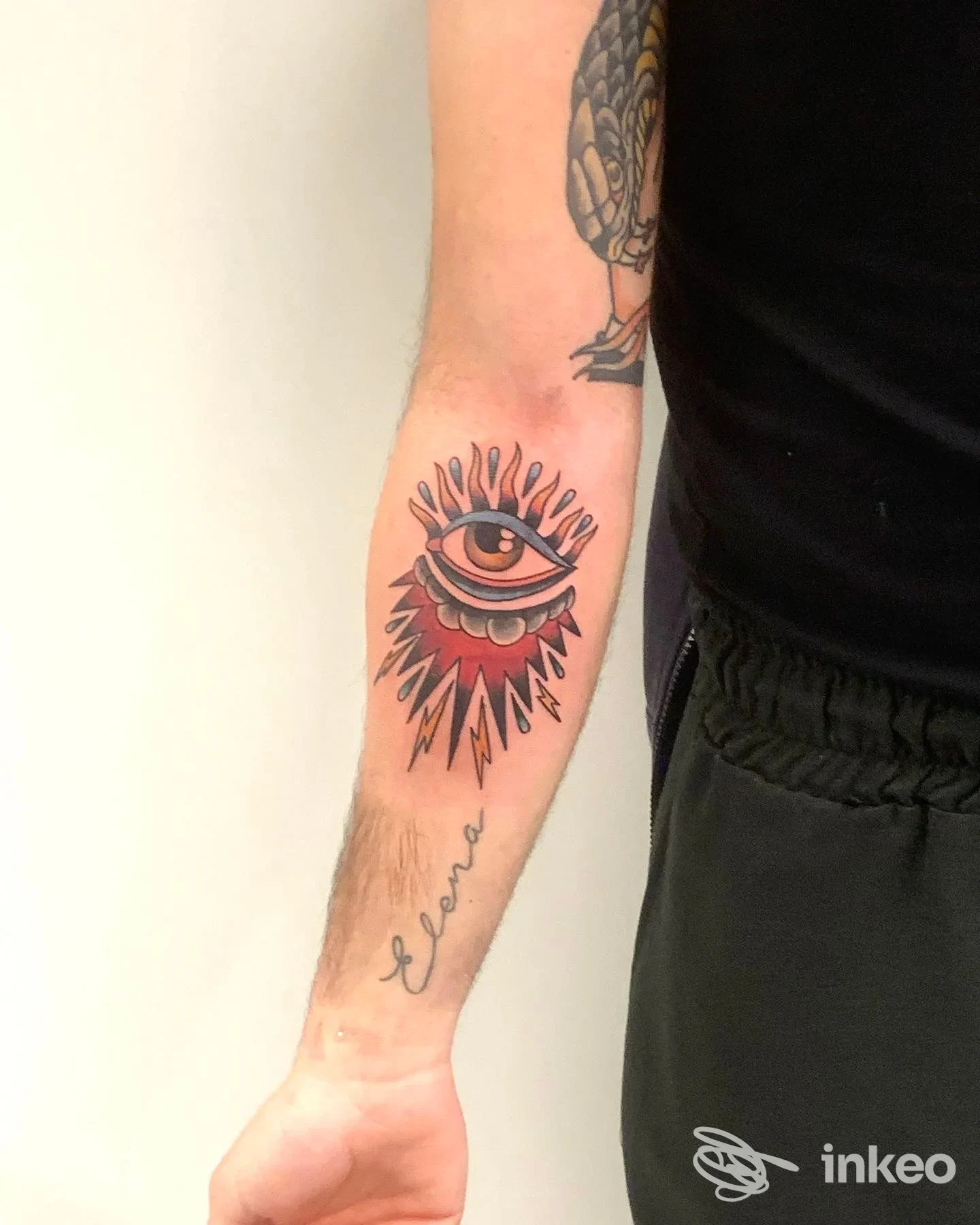 Tatuaggio visibile sull'avambraccio raffigurante un occhio al centro con raggi rossi e neri, realizzato direttamente sulla pelle, sfondo chiaro.