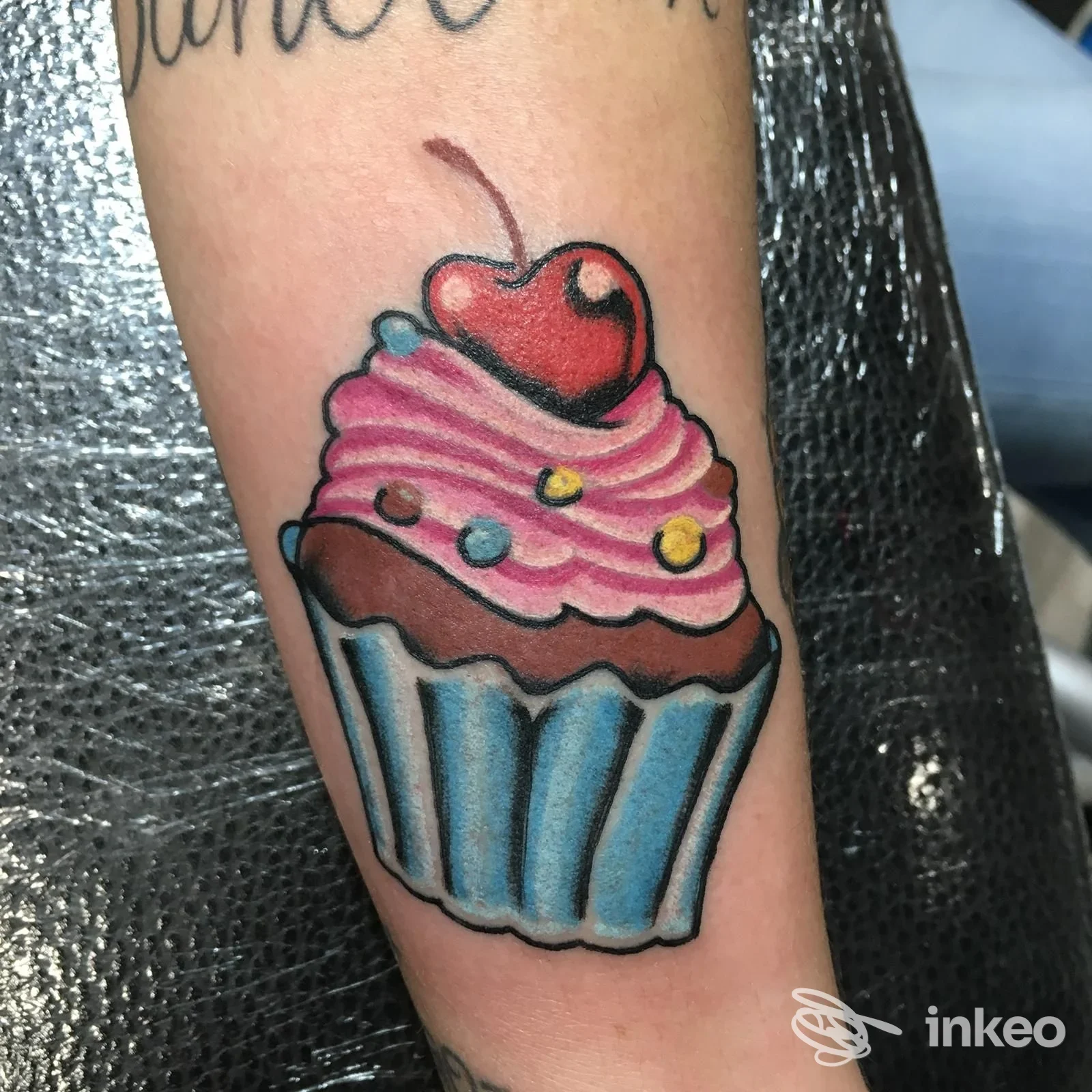 Tatuaggio su pelle raffigurante una cupcake rosa con glassa, ciliegia sopra e dettagli colorati sull’avambraccio.