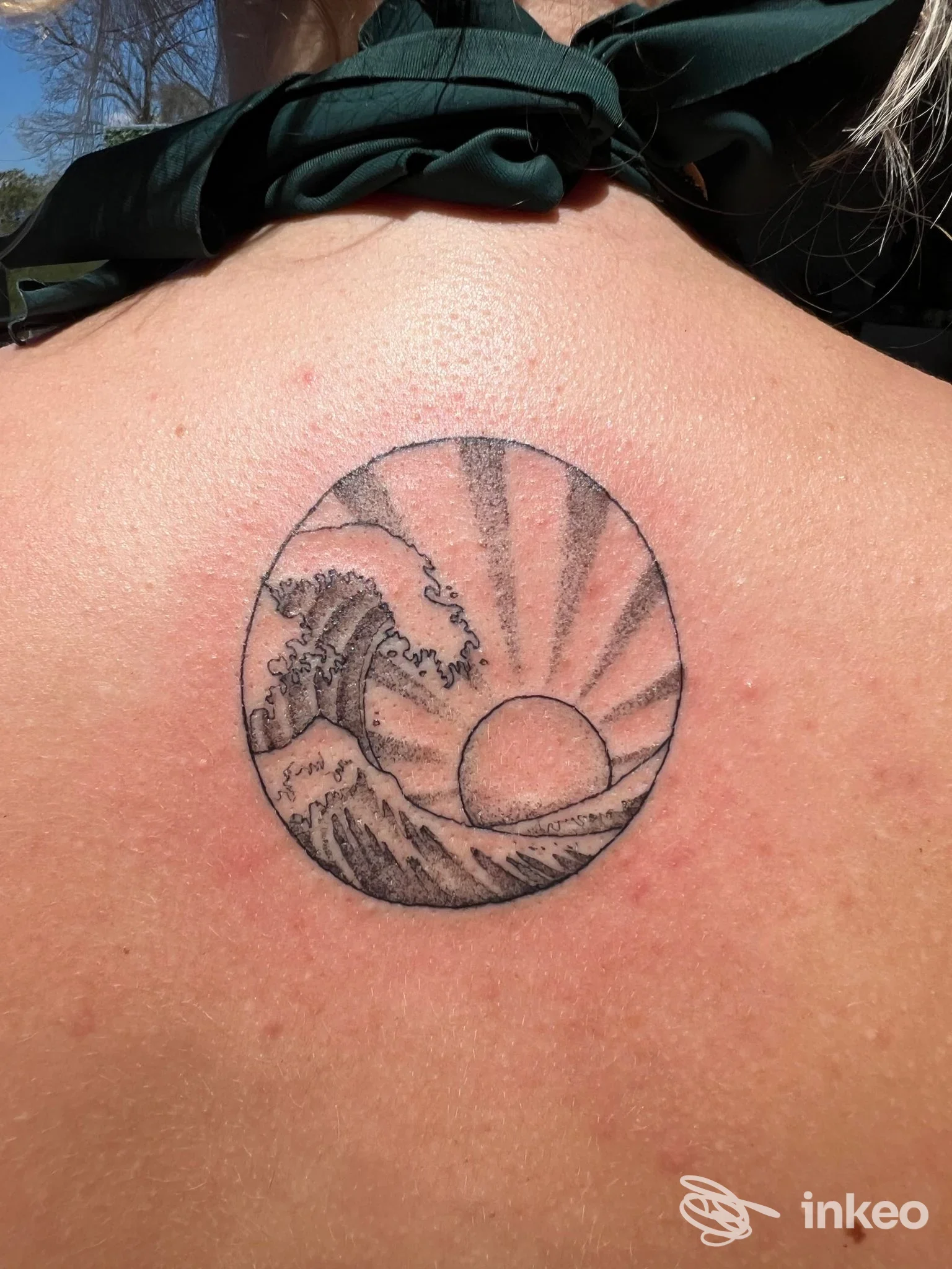 Tattoo circolare con onda e sole all'orizzonte sulla spalla, eseguito in nero.