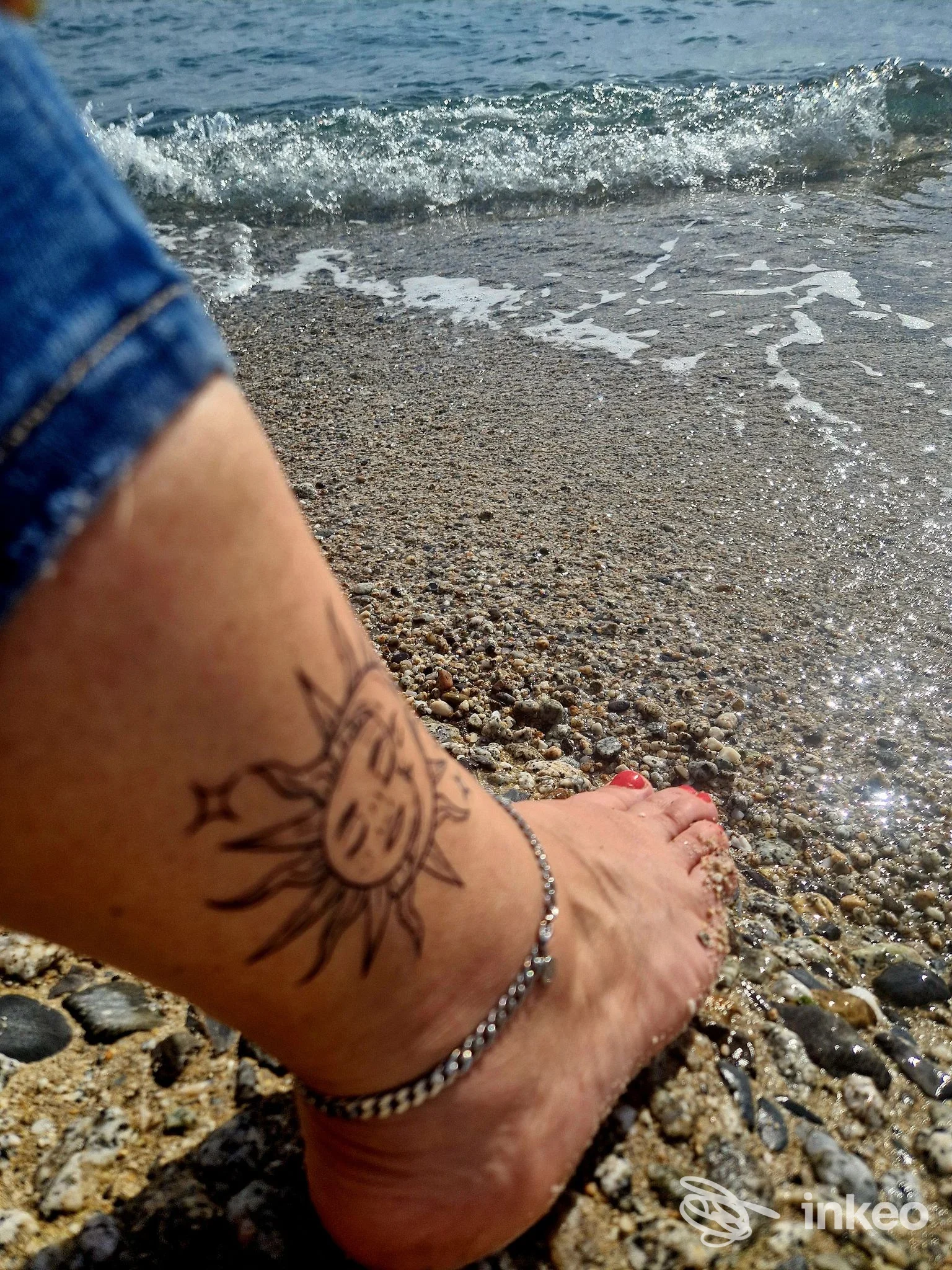 Parte inferiore della gamba con tatuaggio a sole stilizzato, indossando una cavigliera, su spiaggia di ciottoli con mare sullo sfondo.