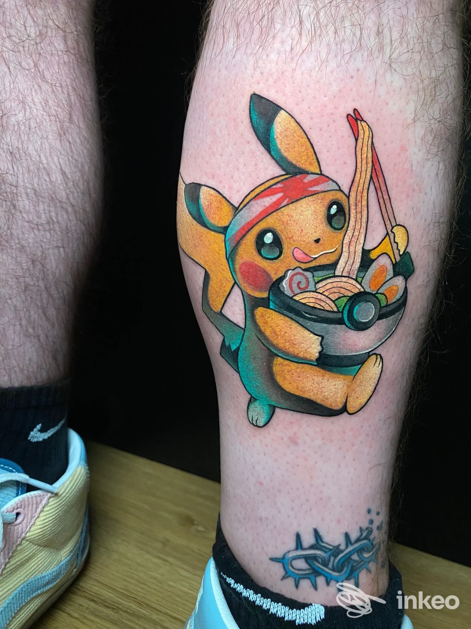 Tattoo su polpaccio raffigurante un Pokemon tipo Eevee in stile cartone animato che mangia ramen, con tonde tonalità gialle, arancioni e verdi; piccolo tatuaggio aggiuntivo blu sotto al polpaccio.