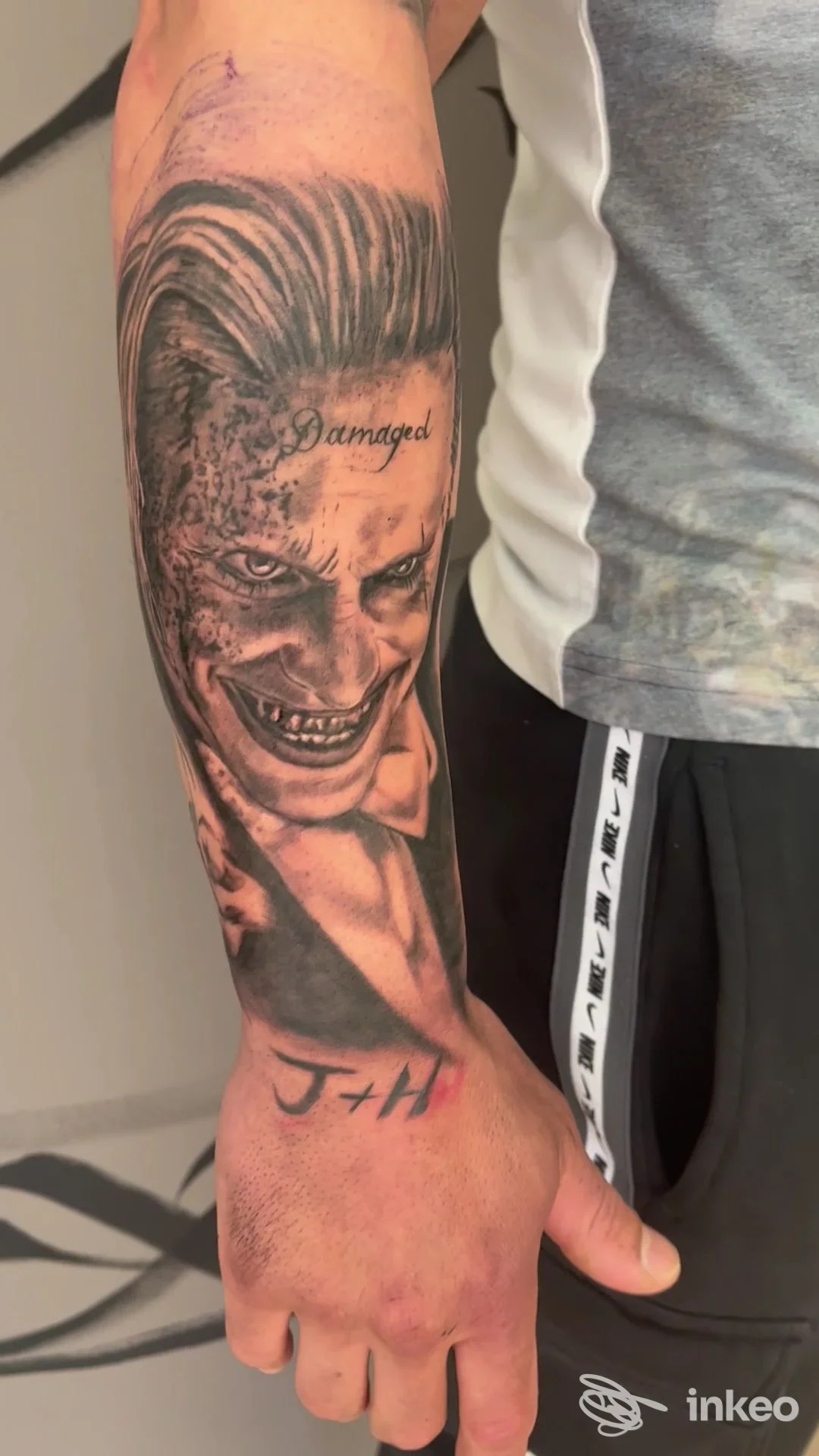 Braccio di una persona con un tattoo realistico a tema Joker sul avambraccio, firma Damaged vicino all'orecchio e iniziali JH sul dorso, realizzato da Fil Tattoo Studio.