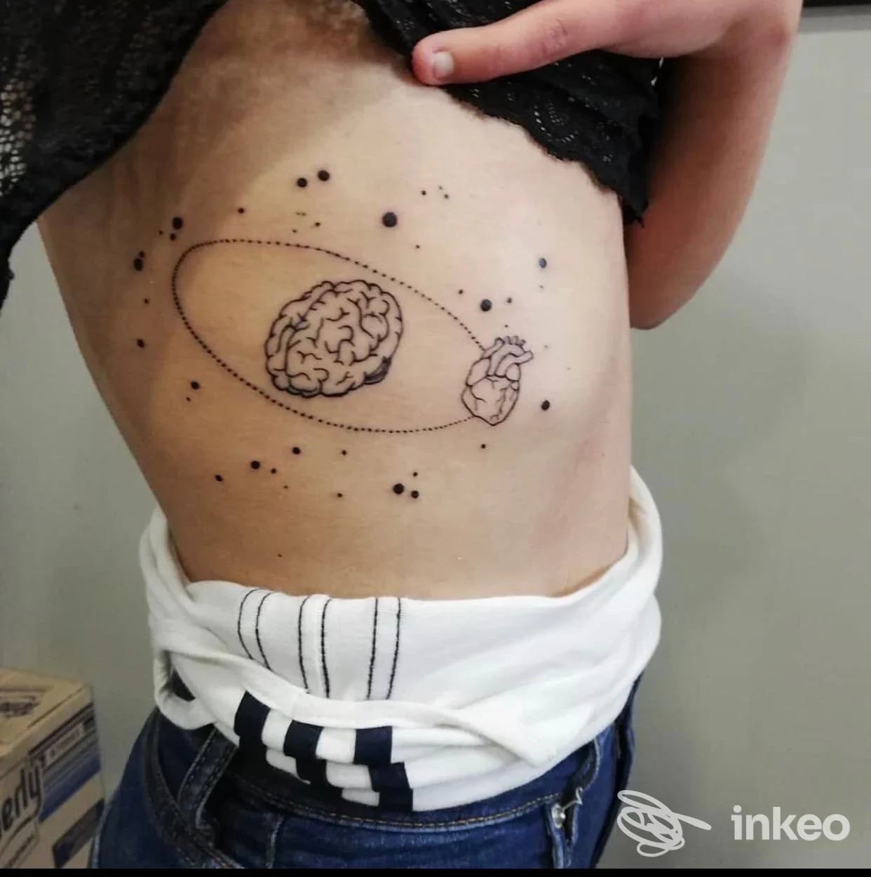 Tatuaggio corporeo visibile sul fianco sinistro: disegno lineare con cervello al centro di un'orbita, un cuore stilizzato e puntini sparsi nello spazio; stile minimale, pelle nuda visibile.