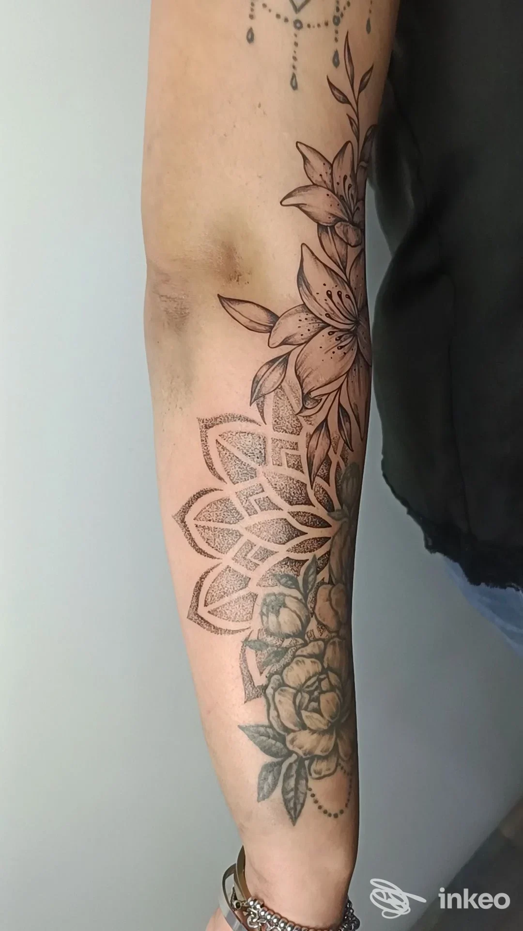Avambraccio con tattoo estetico in stile floreale e mandala, tratteggiature nere e sfumature grigie visibili lungo il braccio; parte della manica è presente, nessun volto visibile.