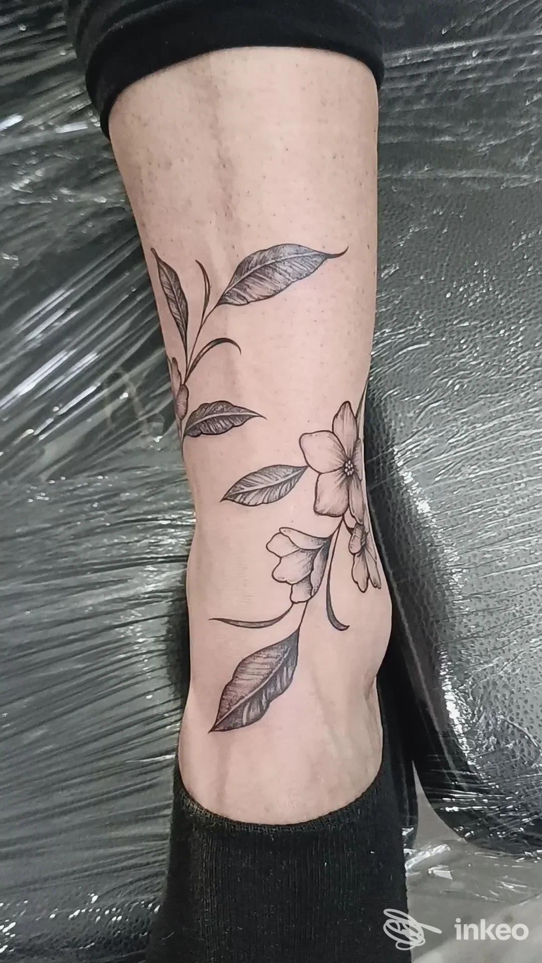 Tattoo floreale sul polpaccio di una persona: linee sottili, foglie e fiori in tinta nera su pelle chiara, con sfondo di tessuto protettivo.
