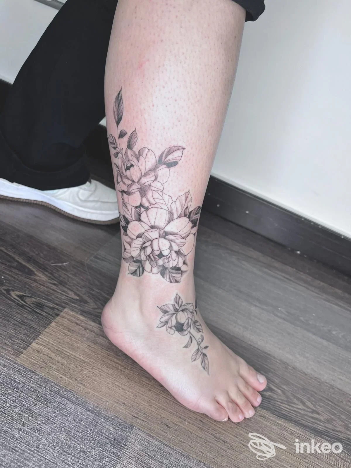 Tattoo floreale in bianco-nero lungo la gamba, partendo dalla caviglia e risalendo verso il polpaccio, eseguito sulla pelle reale.