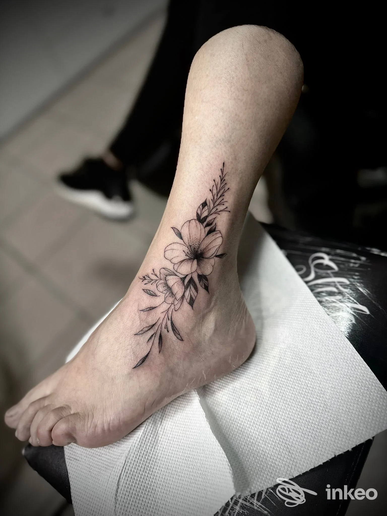 Tattoo floreale in nero-grigio lungo la caviglia e sul dorso del piede, con dettagli di fiori e foglie su pelle reale, stile sottile e delicato.