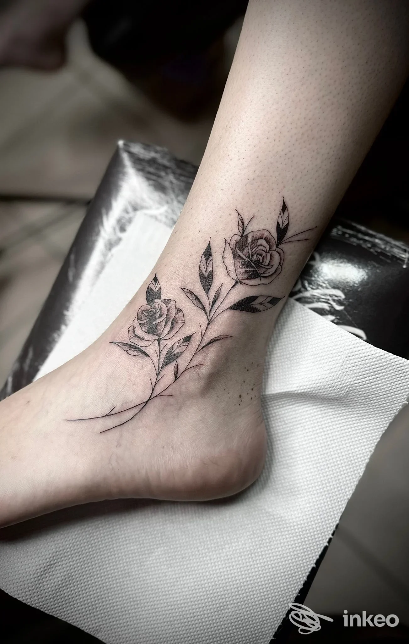 Tatuaggio floreale in tonalità nere/grigie lungo la caviglia e sul piede, stile lineare realistico con foglie e boccioli.